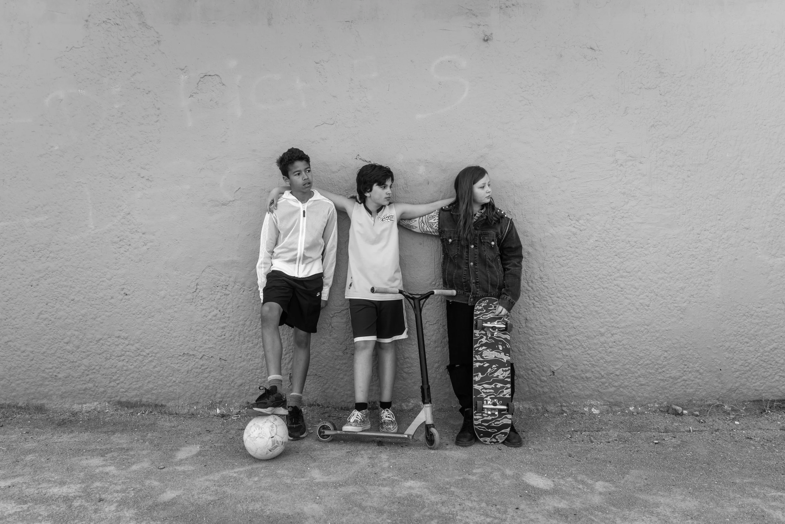 Trois enfants debout contre un mur, un porte un ballon de football, un autre un skateboard, le troisième un vélo ZigZag, en noir et blanc.