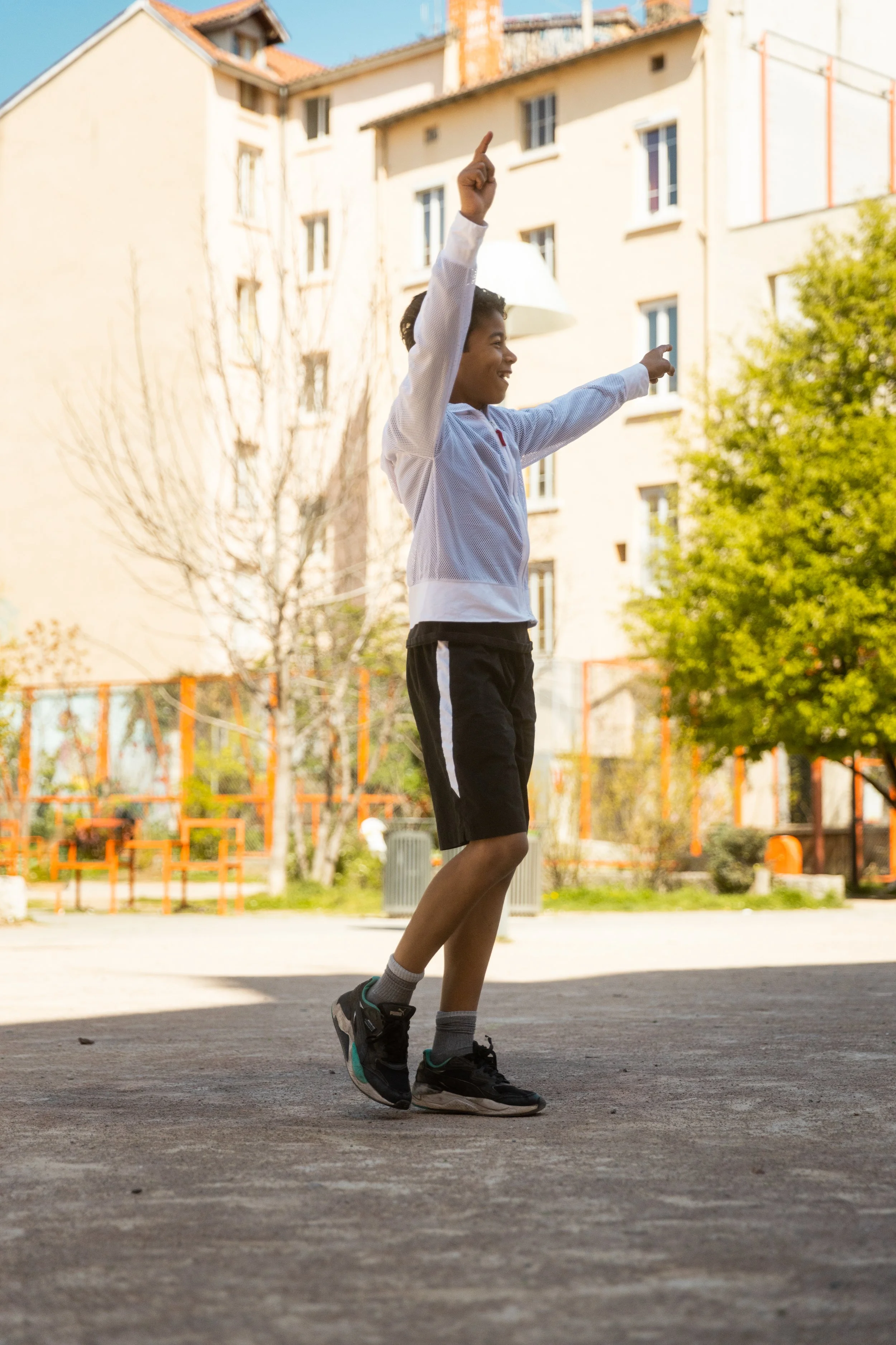 Un garçon jeune en t-shirt et shorts noirs, faisant du saut dans une cour extérieure ensoleillée, avec des immeubles et des arbres en arrière-plan.