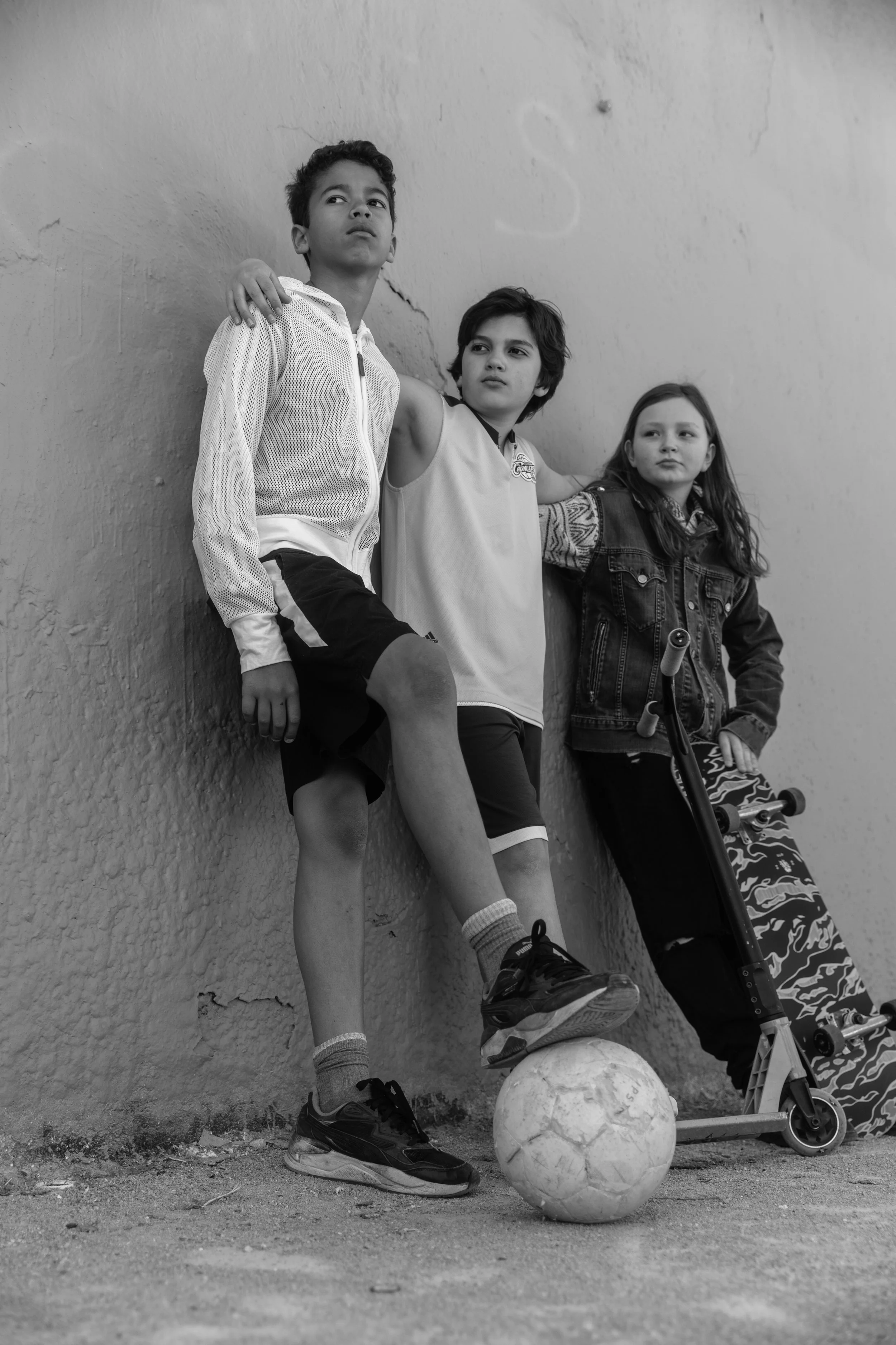 Trois enfants posant contre un mur, avec un ballon de football, un skateboard, et une trottinette.