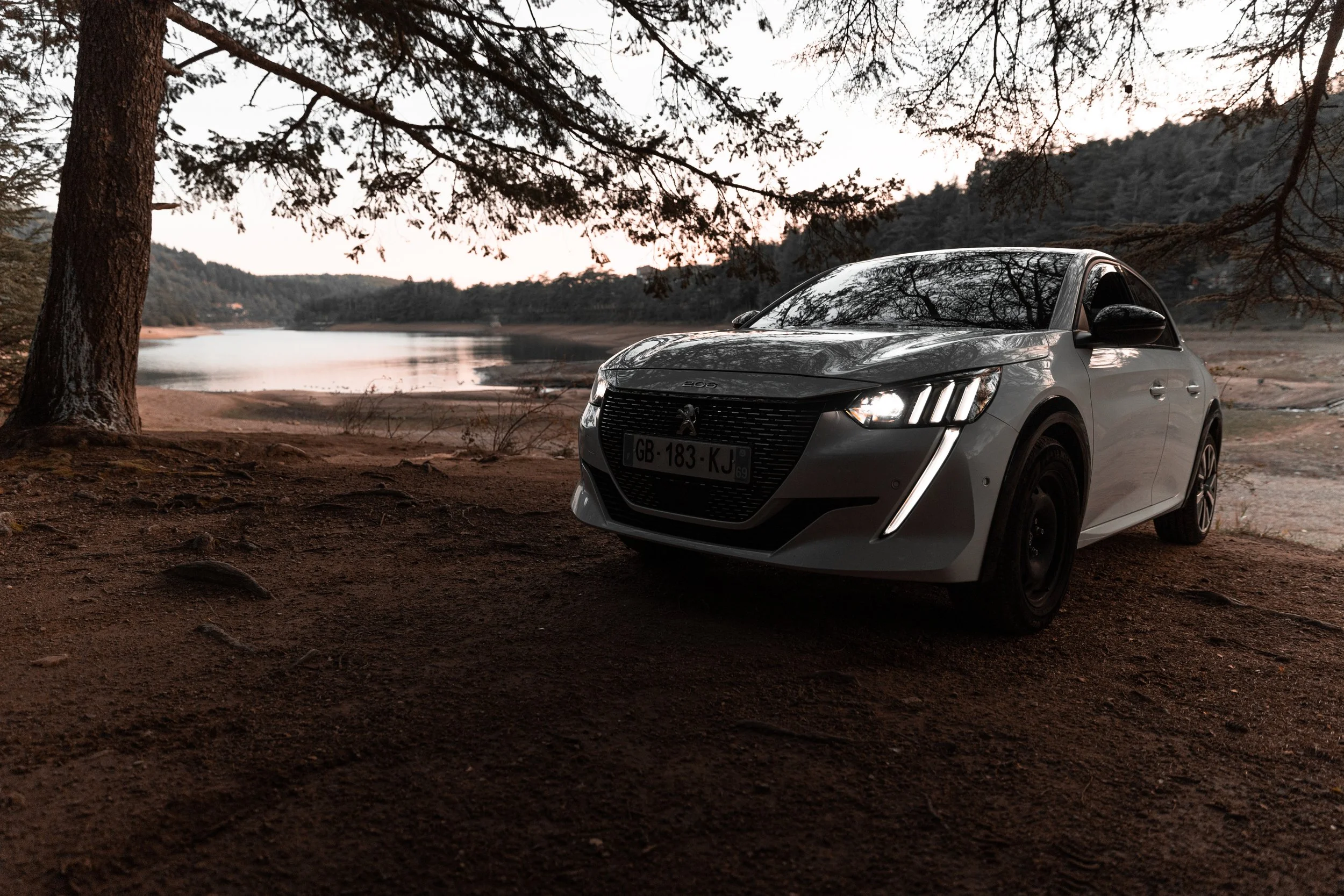 Une voiture blanche garée près d'un lac entouré d'arbres, au crépuscule.