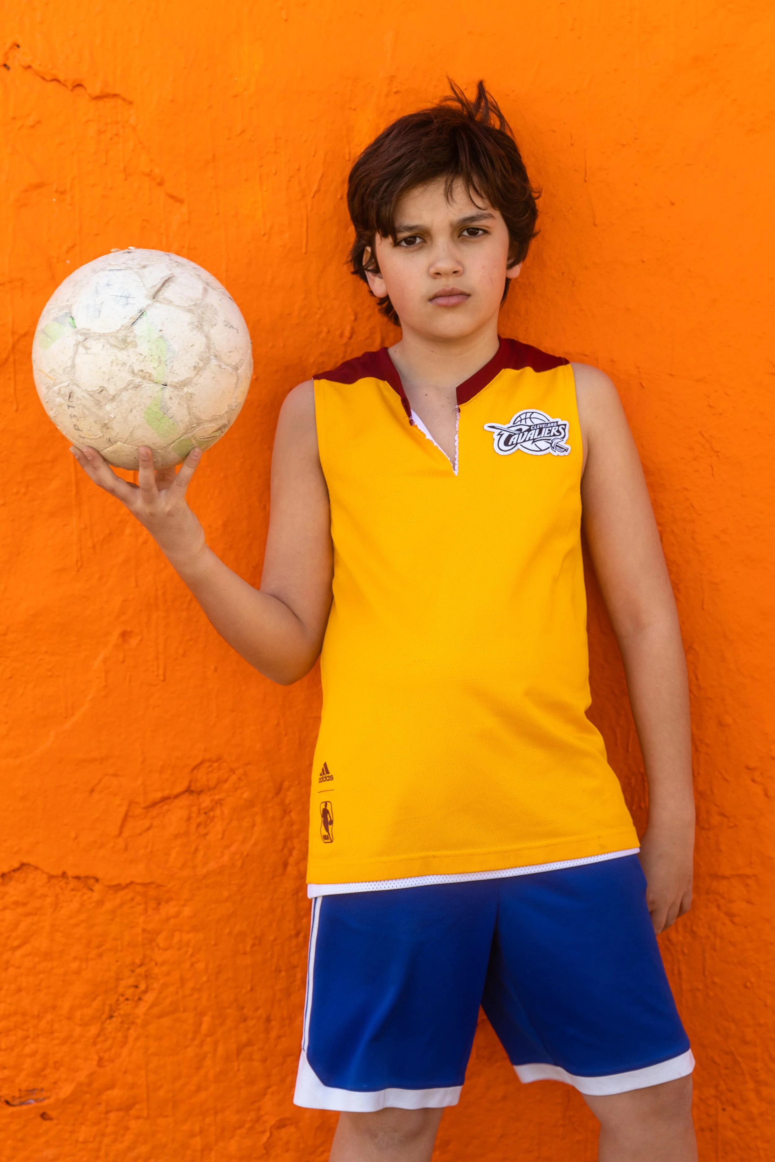 Un garçon avec un maillot de basket jaune et un short bleu, tenant un ballon de basketball face à un mur orange.
