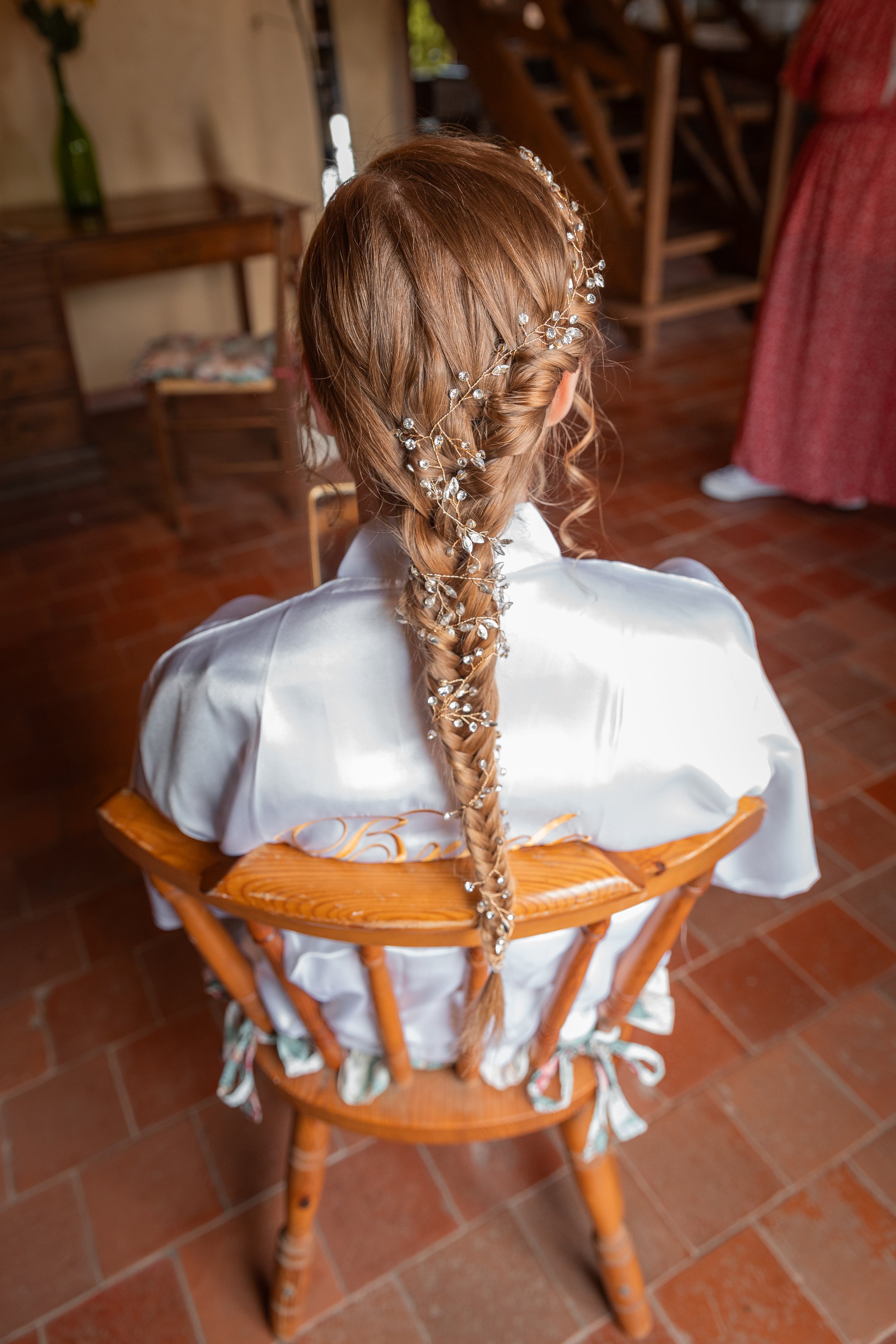 Une femme avec de longs cheveux roux coiffés en tresse ornée de perles et de fil métallique, vue de dos, assise sur une chaise en bois, portant une robe blanche. Le décor intérieur est chaud et rustique, avec un sol en tuiles et des meubles en bois.