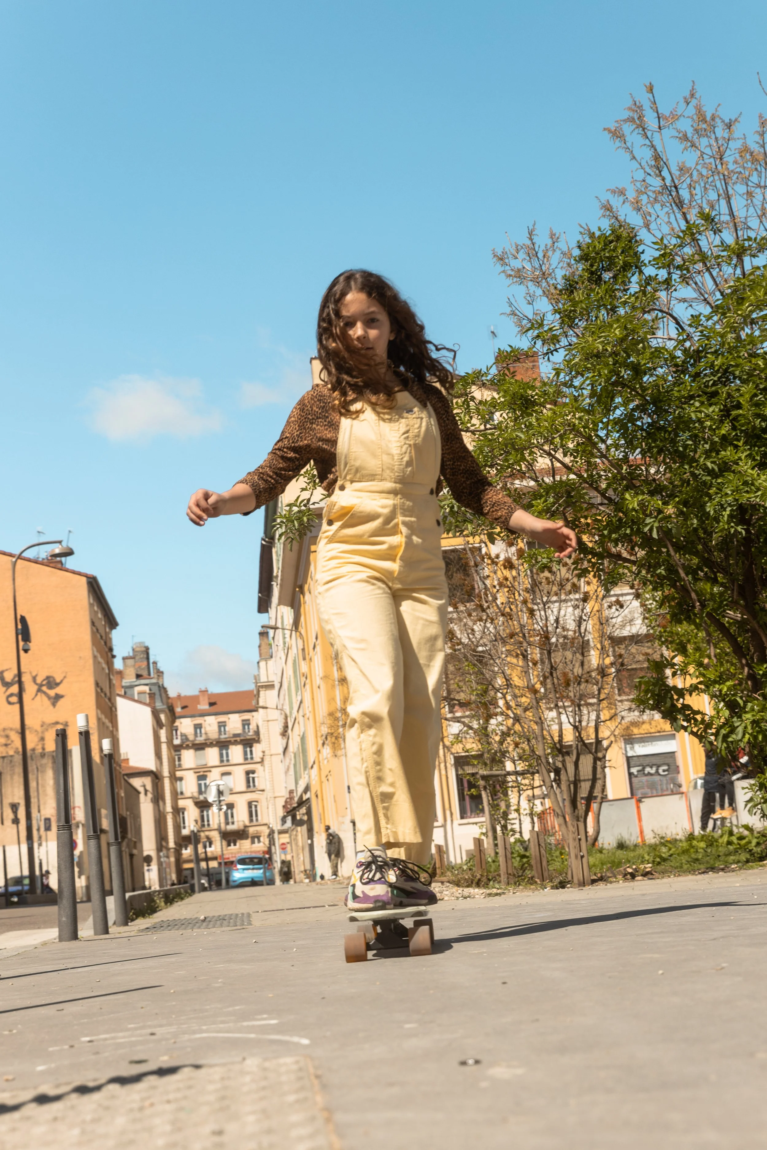 Fille faisant du skateboard dans une rue urbaine ensoleillée, avec des bâtiments résidentiels en arrière-plan, portant un ensemble beige et des chaussures colorées.