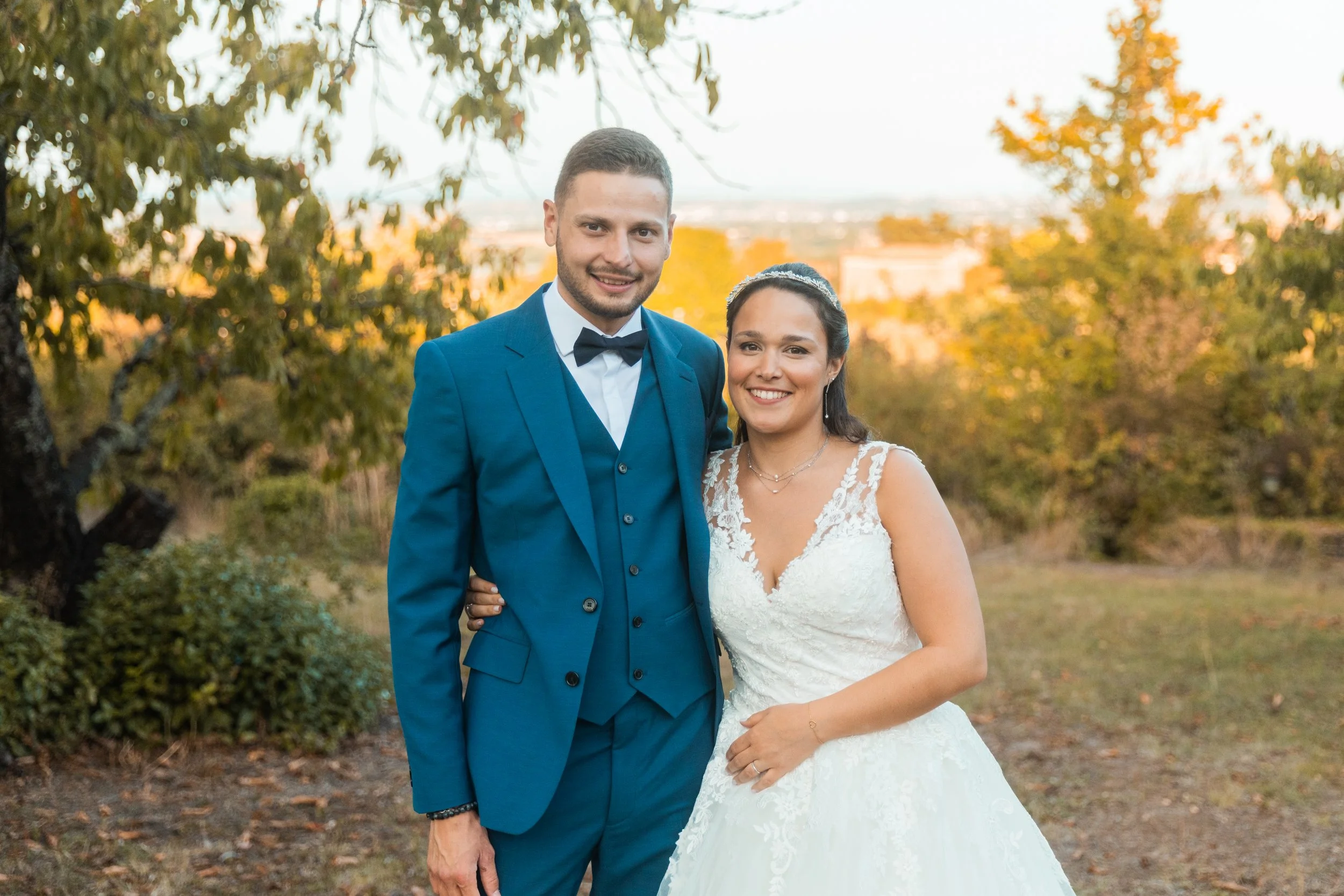 Un couple habillé en costume de mariage, debout dans un parc avec des arbres aux feuilles automnales, souriant à la caméra, lors d'une cérémonie de mariage en plein air.