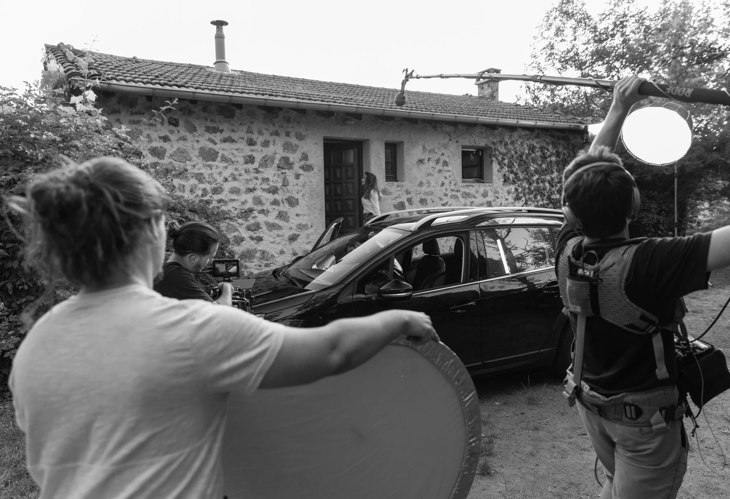 Tournage d'une scène filmée devant une maison en pierre, avec une voiture noire en premier plan, une femme debout devant la maison, et deux personnes avec du matériel de tournage.