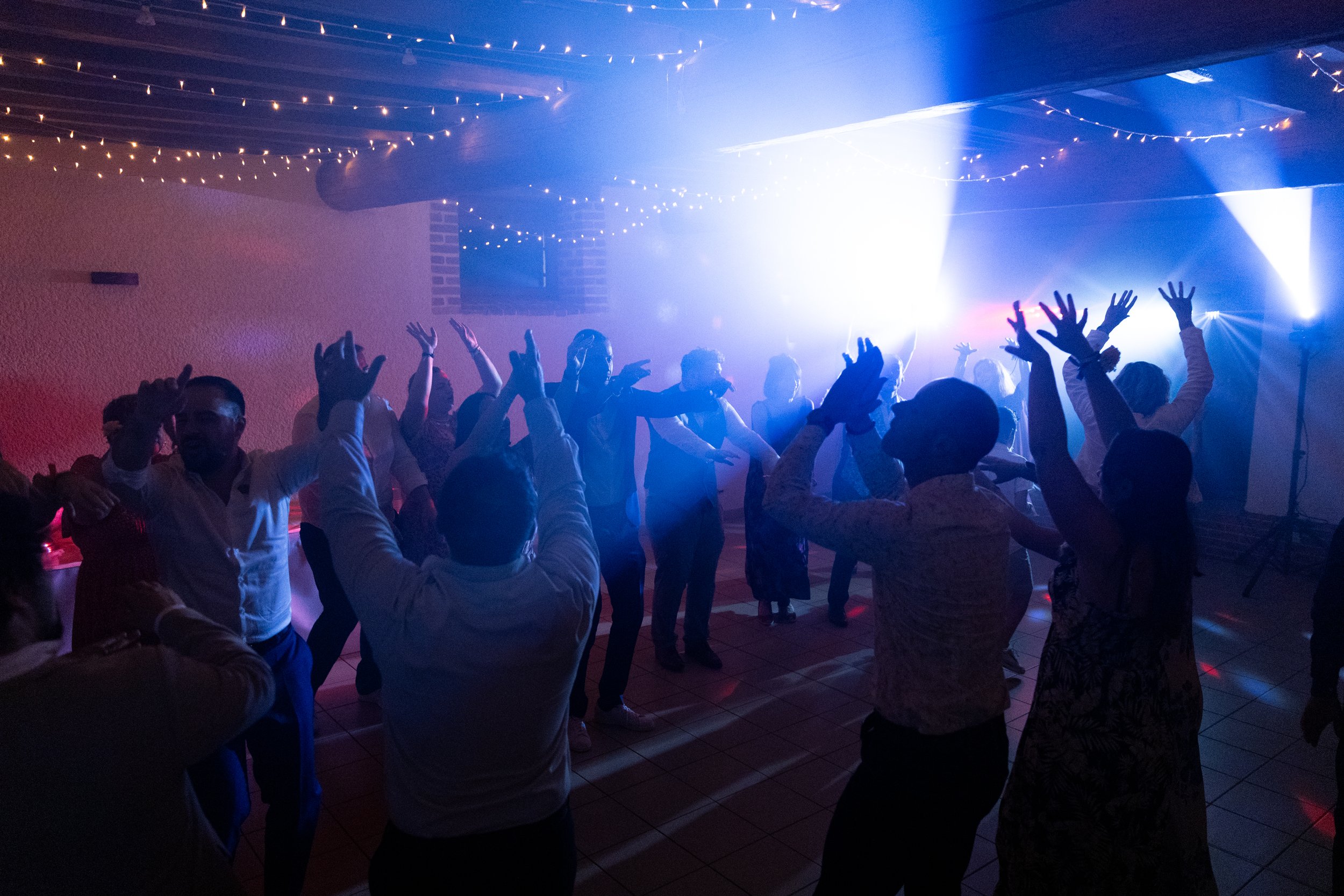 Groupe de personnes dansant lors d'une fête avec des lumières colorées et guirlandes lumineuses suspendues au plafond.