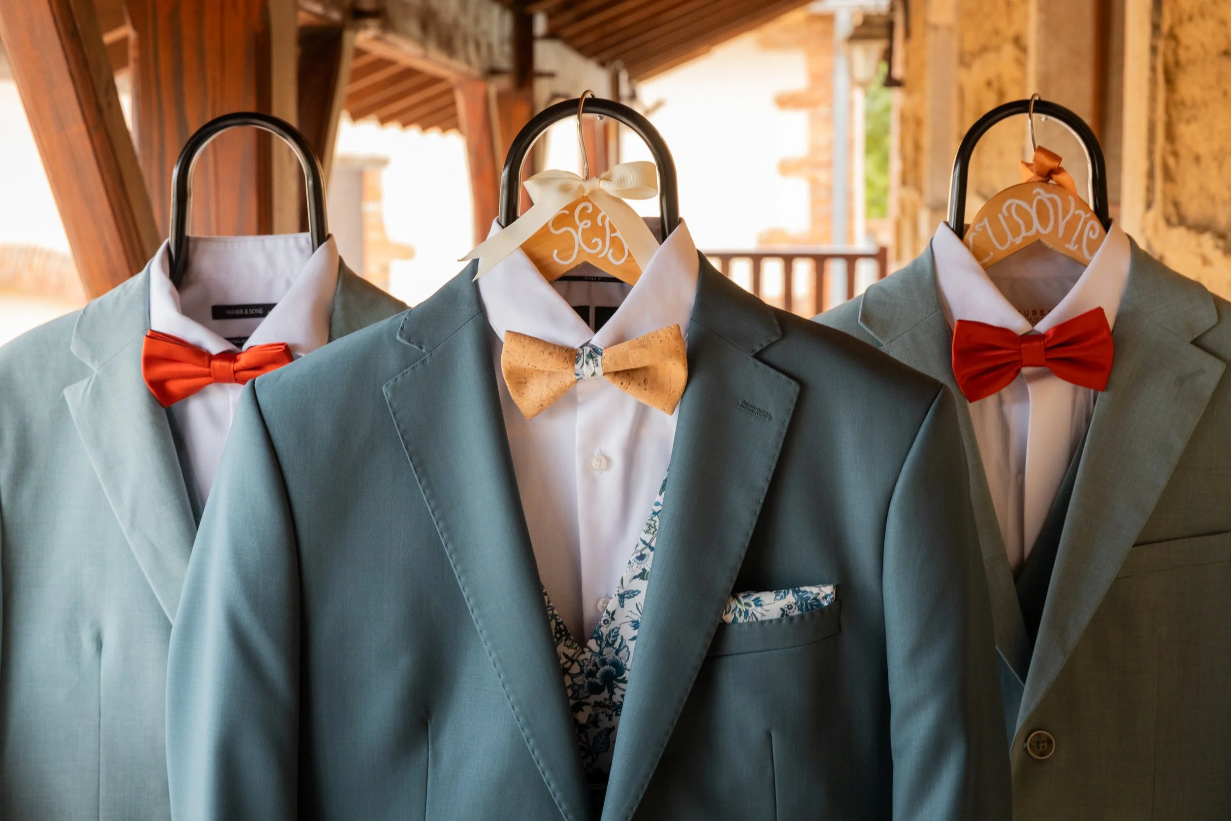 Costumes de mariage avec vestes gris clair, chemises blanches et nœuds papillons rouges, suspendus sur des cintres en bois dans une ambiance chaleureuse.