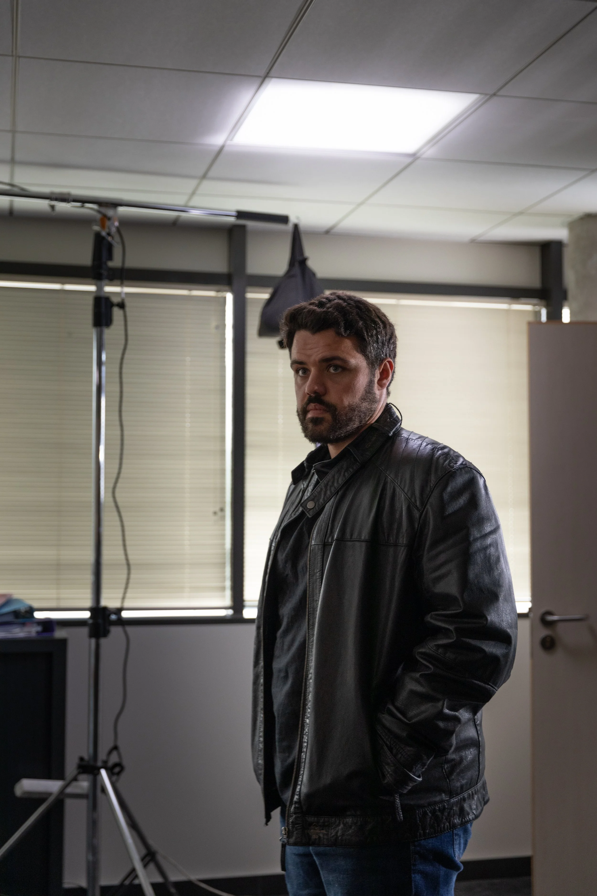 Un homme avec une barbe portant une veste en cuir noir, dans une pièce avec des fenêtres à stores, un plafond suspendu et des équipements de studio.