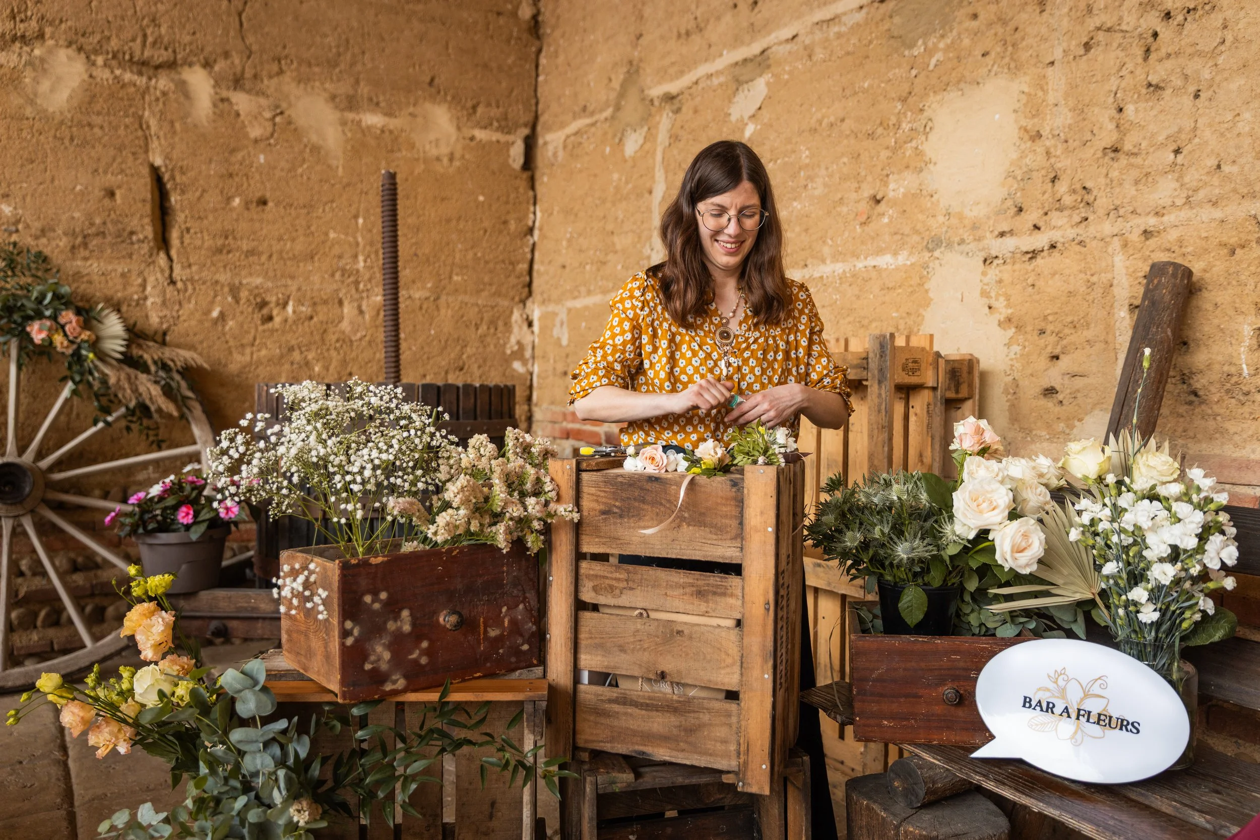Une femme arrangeant des fleurs dans un atelier de fleuriste, avec des fleurs blanches et un panneau "Bar à Fleurs".