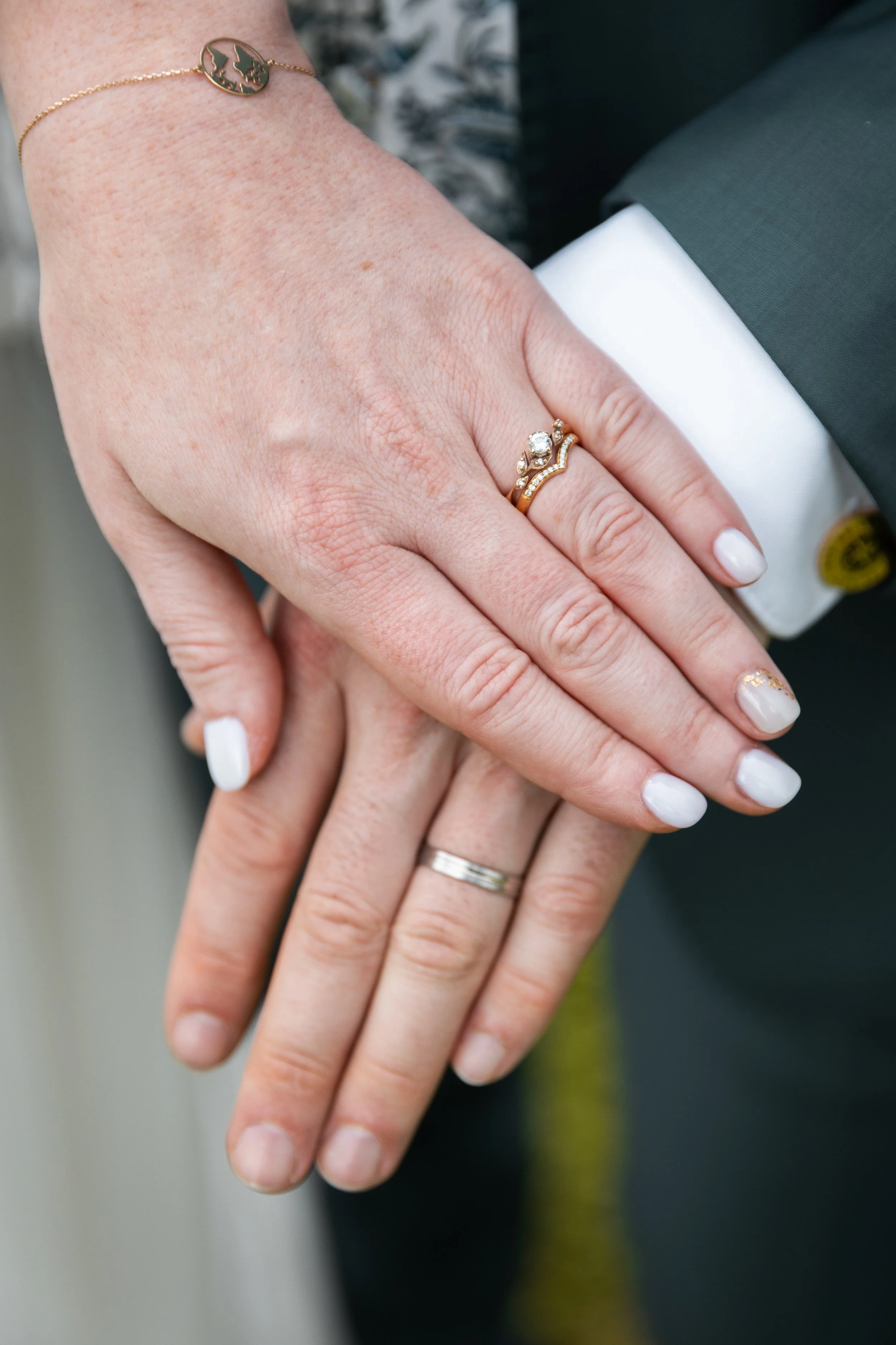 Deux mains entrelacées, portant des alliances, l'une avec des ongles peints en blanc et l'autre avec des ongles décorés de dessins noirs, portant une bague en or avec des diamants, une bracelet en or avec un symbole de deux oiseaux, et un costume ave