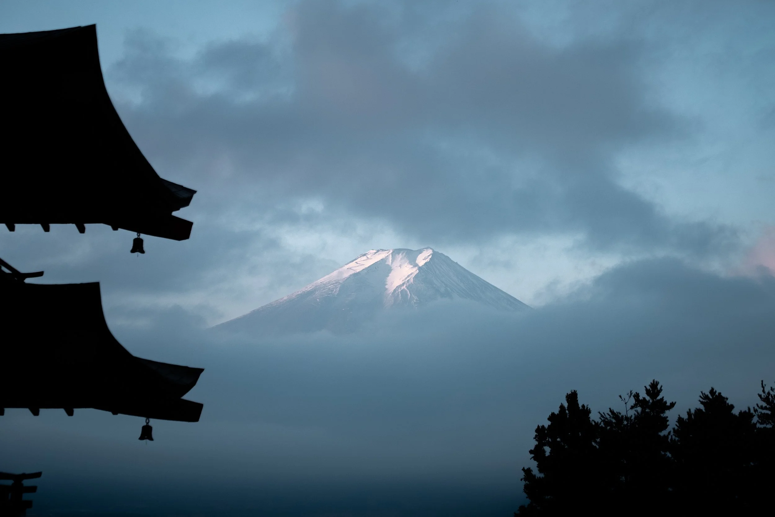 Mont Fuji enneigé vu à travers des nuages, avec des toits traditionnels japonais en silhouette à gauche et des arbres à droite.