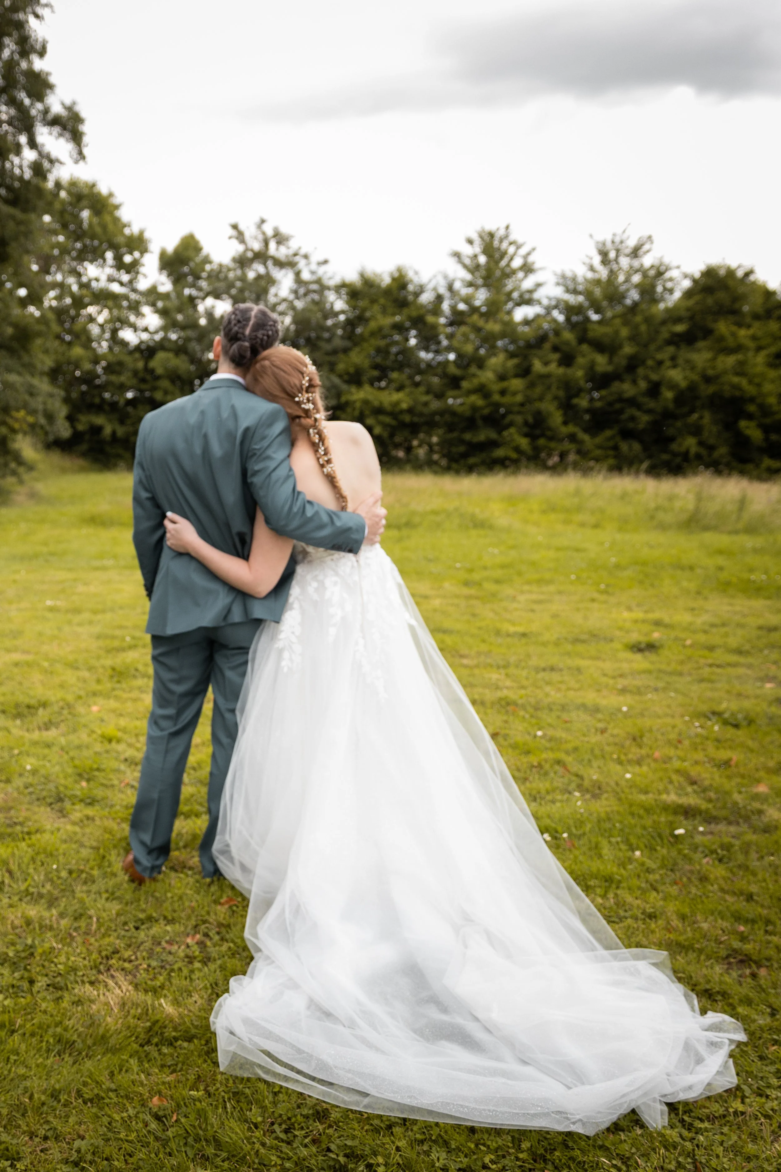 Un couple en habits de mariage, embrassés, vu de dos, dans un champ vert avec des arbres en arrière-plan, ciel nuageux.