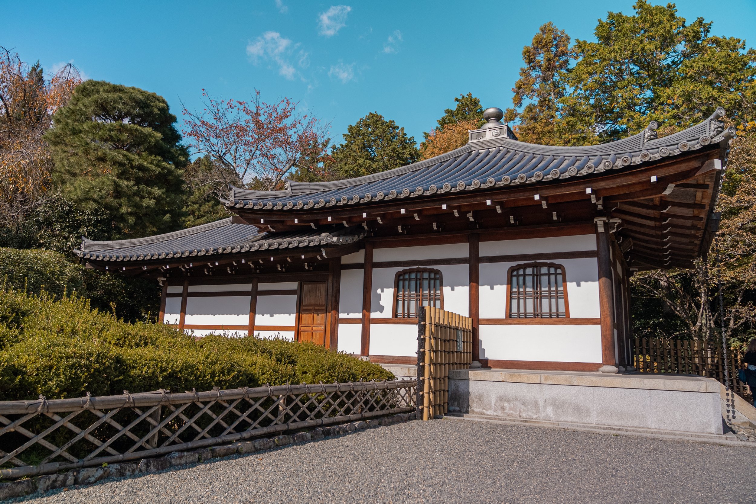 Bâtiment traditionnel japonais avec toit en tuiles noires, murs blancs et fenêtres en bois, entouré d'arbres et de buissons, sous un ciel bleu.