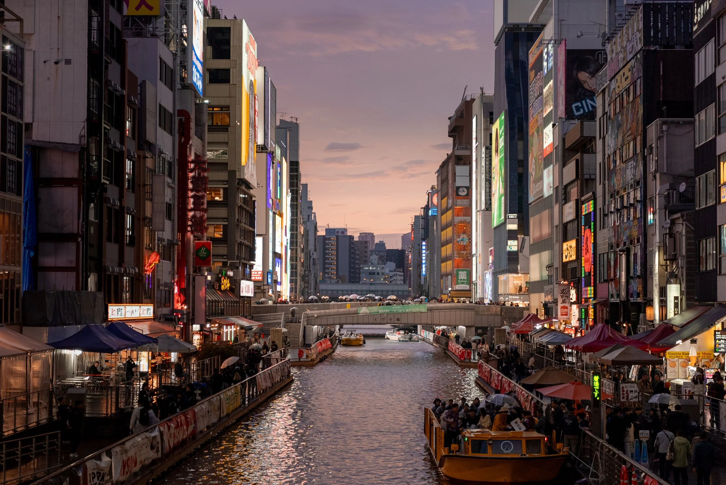 Une vue de nuit d'une ville japonaise avec un canal bordé de bâtiments éclairés, de nombreuses enseignes lumineuses et des passants sous des parapluies.