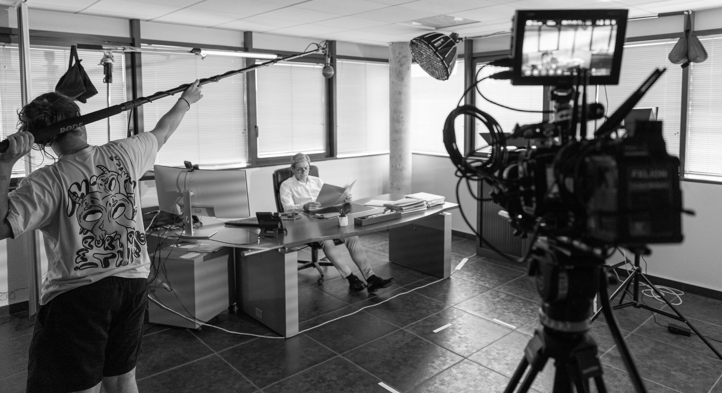 Une personne assise à un bureau, lisant un document, dans un environnement de bureau, avec une caméra et une équipe de tournage en arrière-plan.