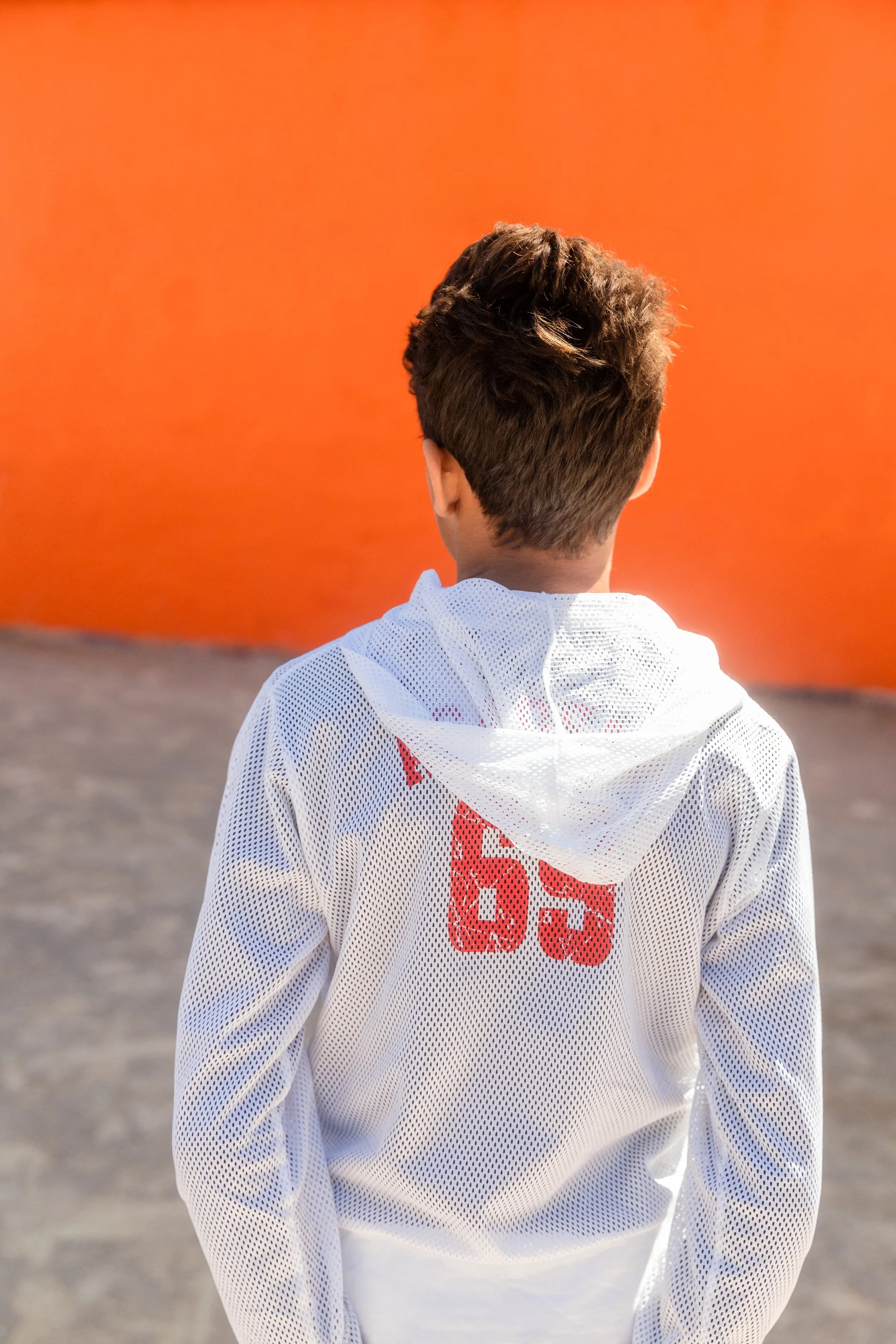 Un garçon vu de dos portant un maillot blanc avec le nombre 55 rouge, se tenant devant un mur orange.