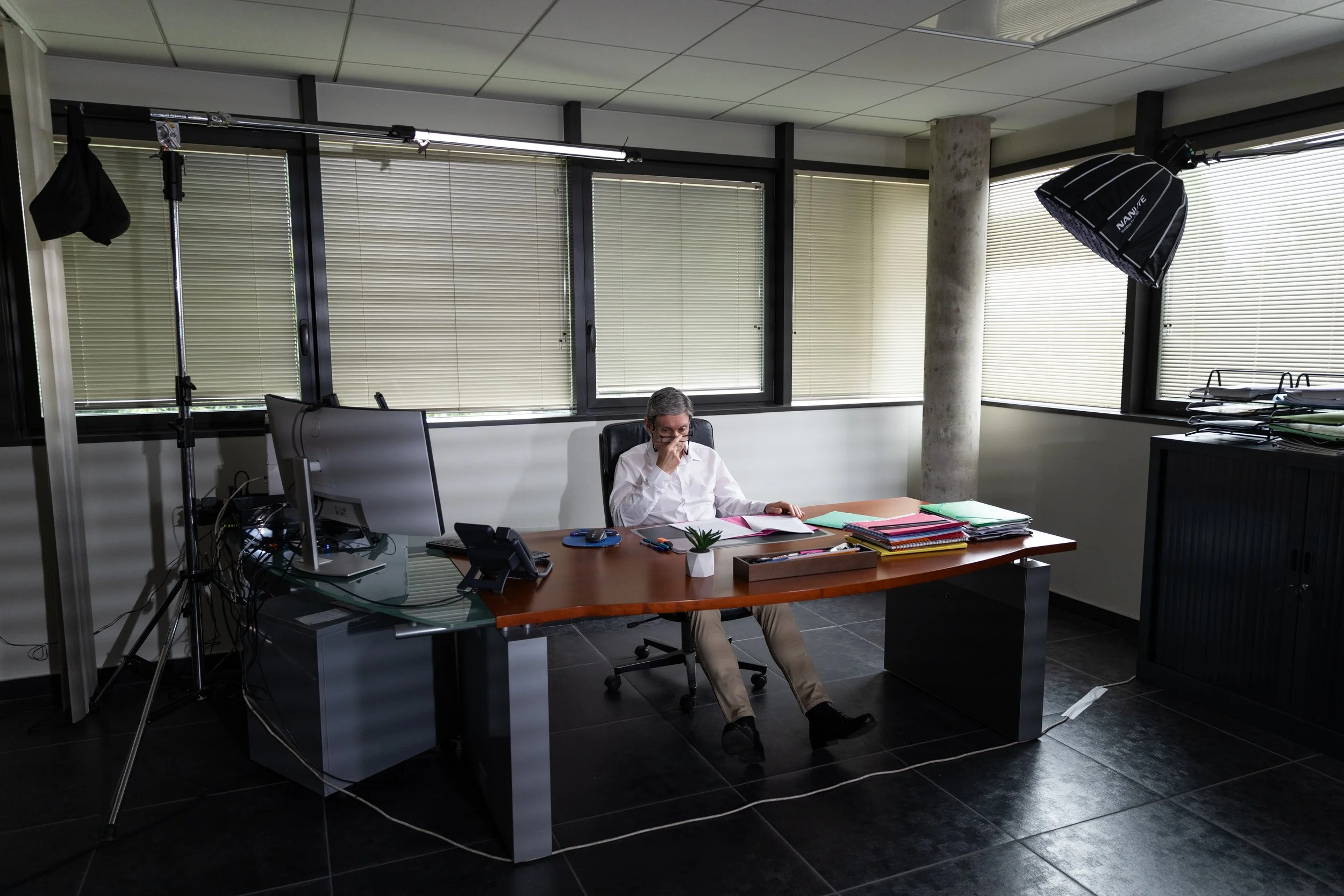 Un homme assis à un bureau avec plusieurs dossiers, une plante en pot, un téléphone, un ordinateur et du matériel de bureau, dans une salle lumineuse avec des grandes fenêtres et des stores vénitiens, installé dans une chaise de bureau.