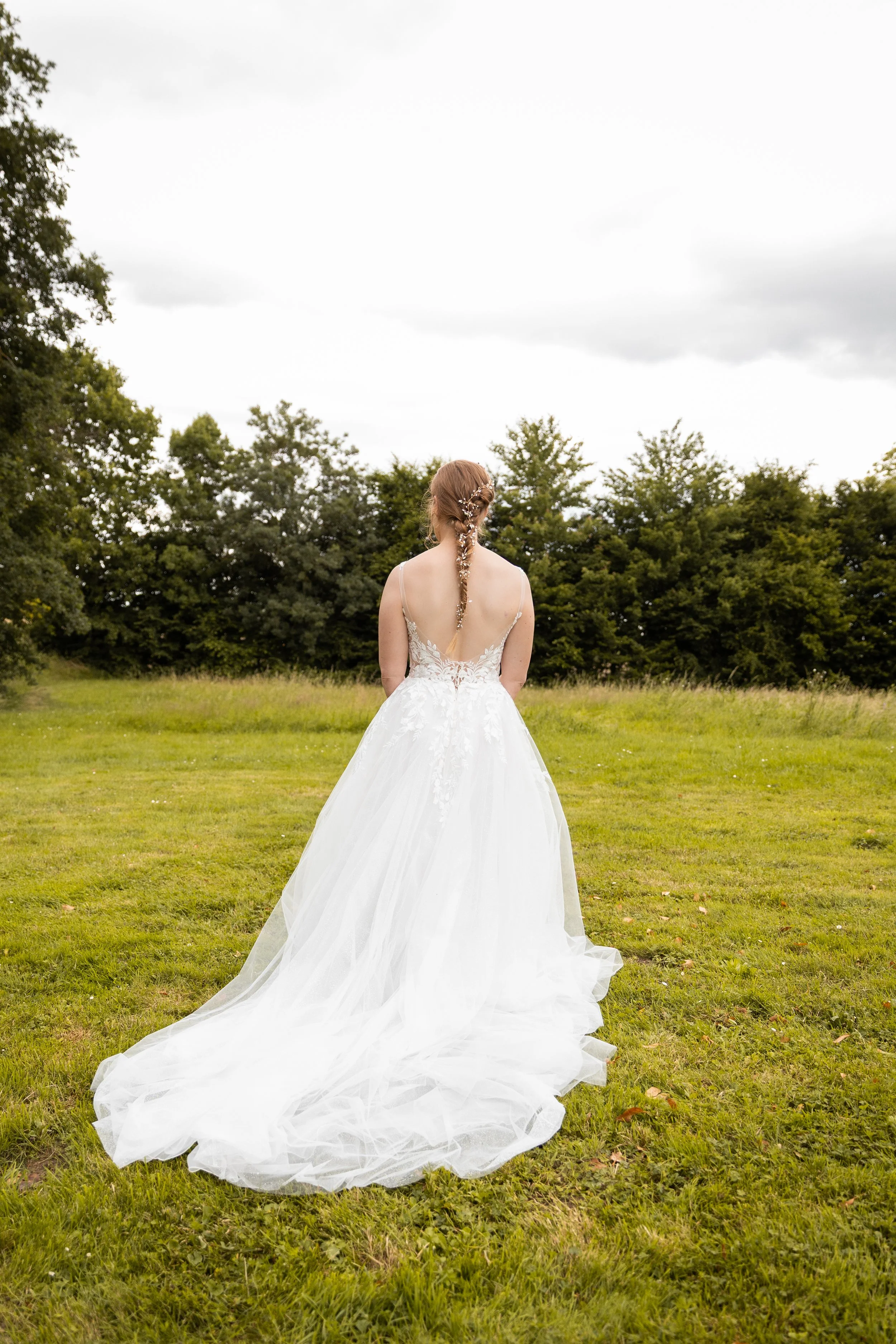 Une femme en robe de mariée blanche dans un champ vert, vue de dos, avec des arbres en arrière-plan et un ciel nuageux.