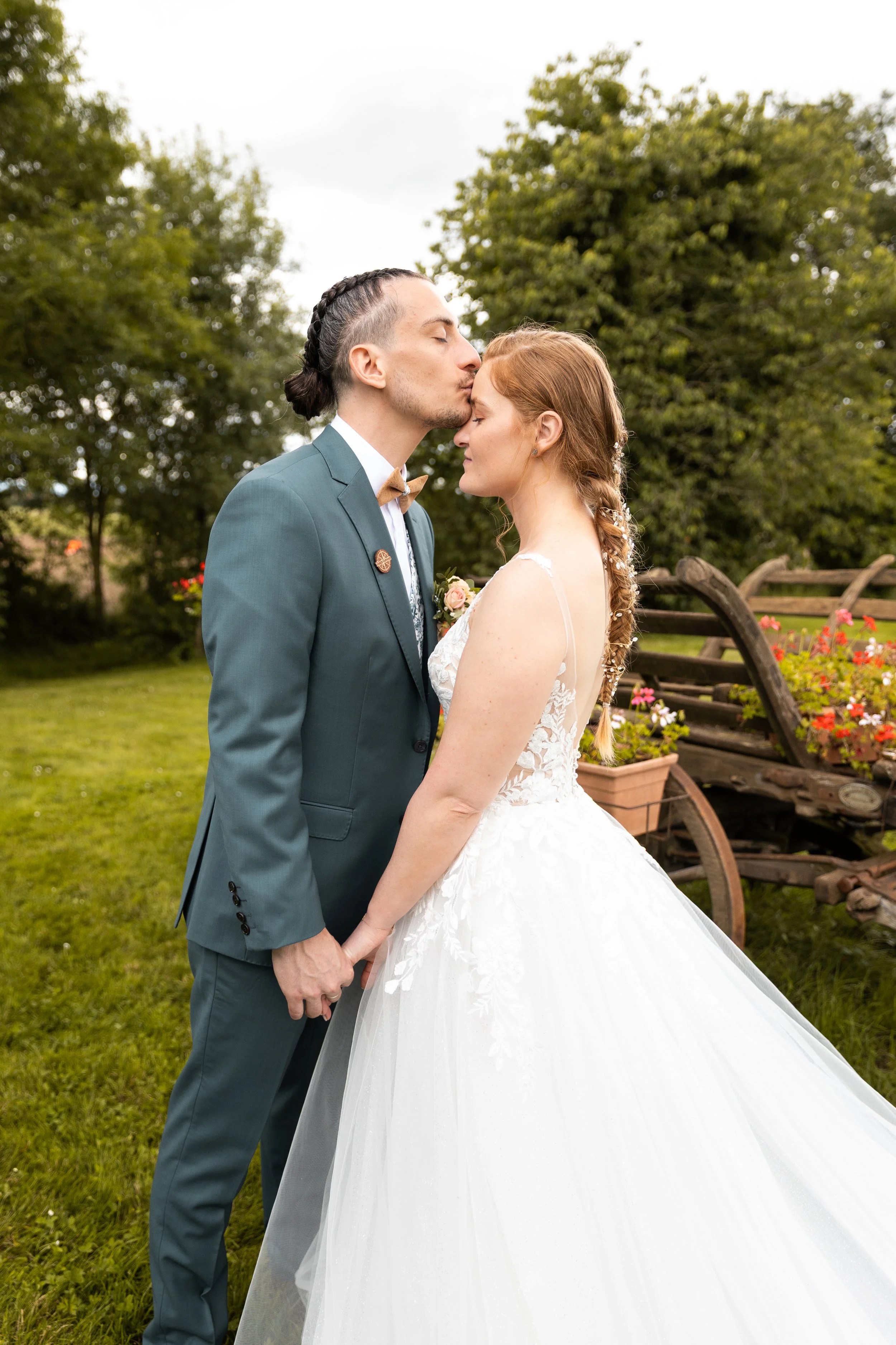 Un couple de mariés en plein baiser dans un jardin verdoyant, entouré de fleurs et de vieux véhicules en bois.