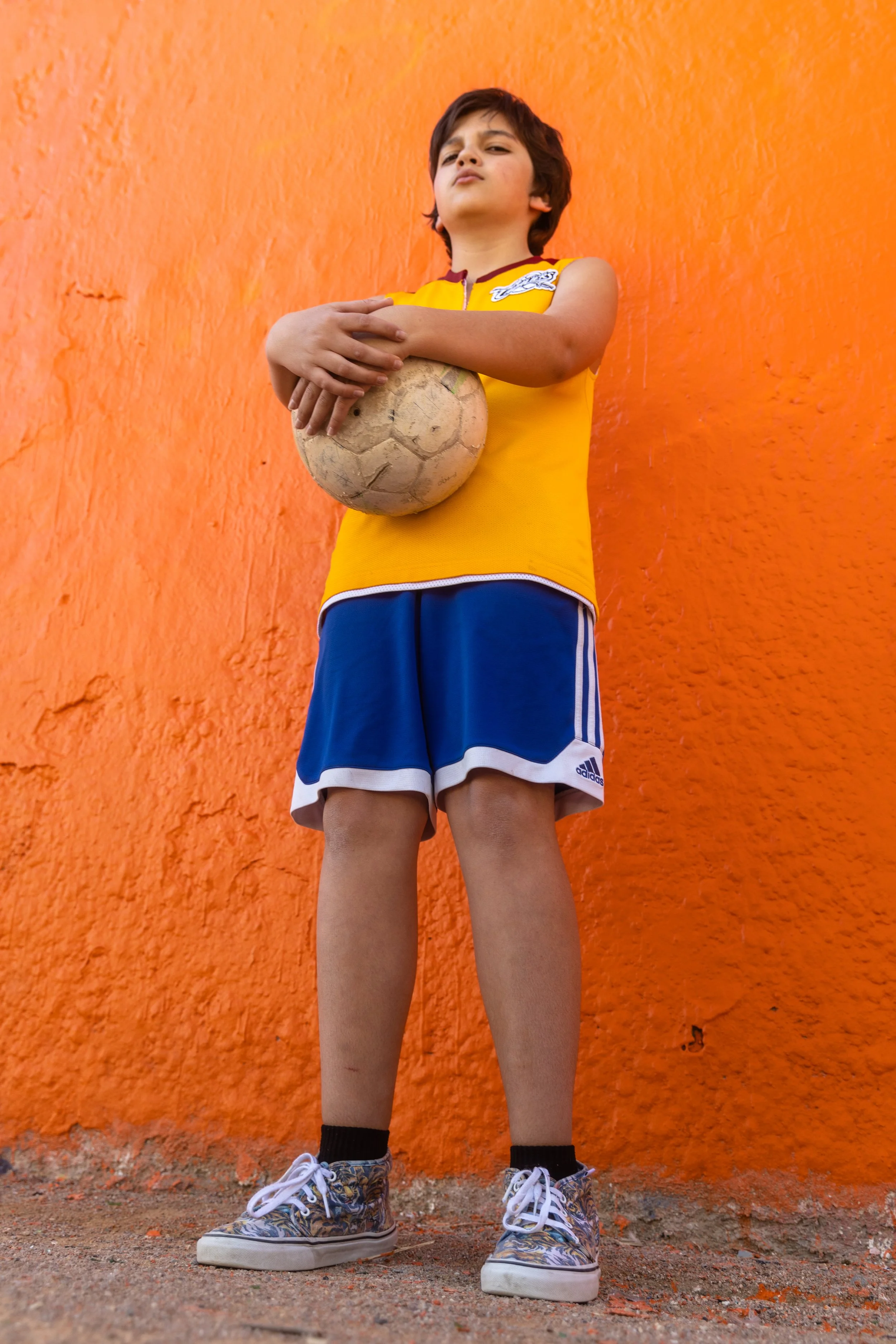 Un garçon debout devant un mur orange, tenant un ballon de football contre lui.