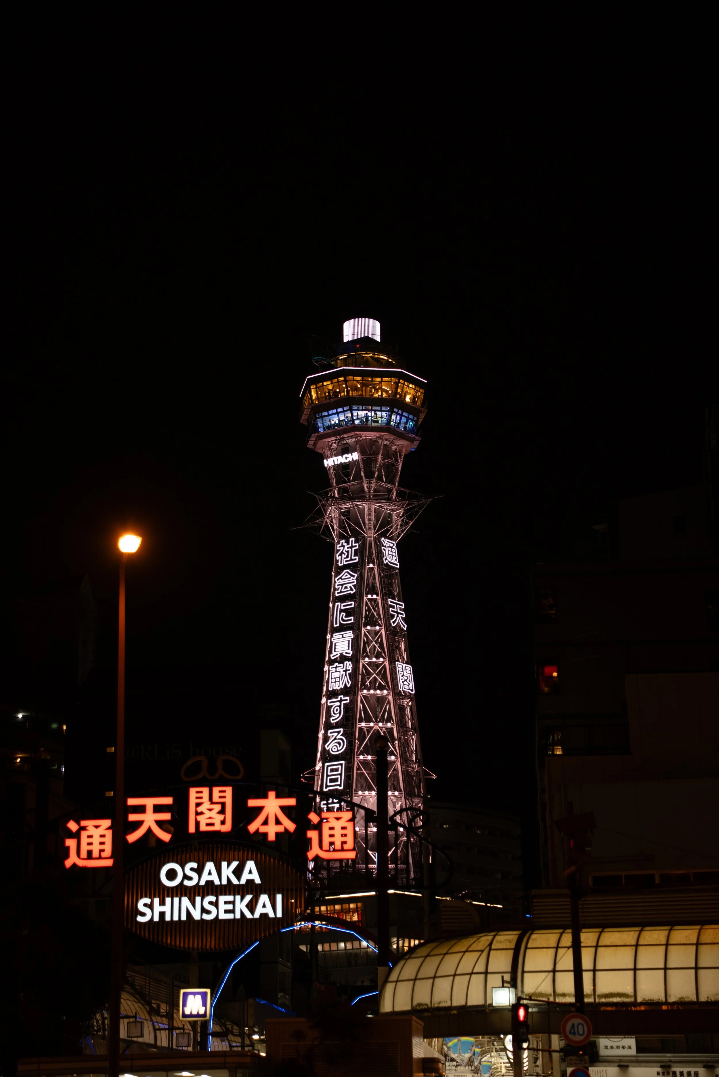 Tour de l'horloge de l'Observatoire à Osaka, illuminée la nuit, avec panneaux en japonais et en anglais indiquant 'Osaka Shinsaibashi'.