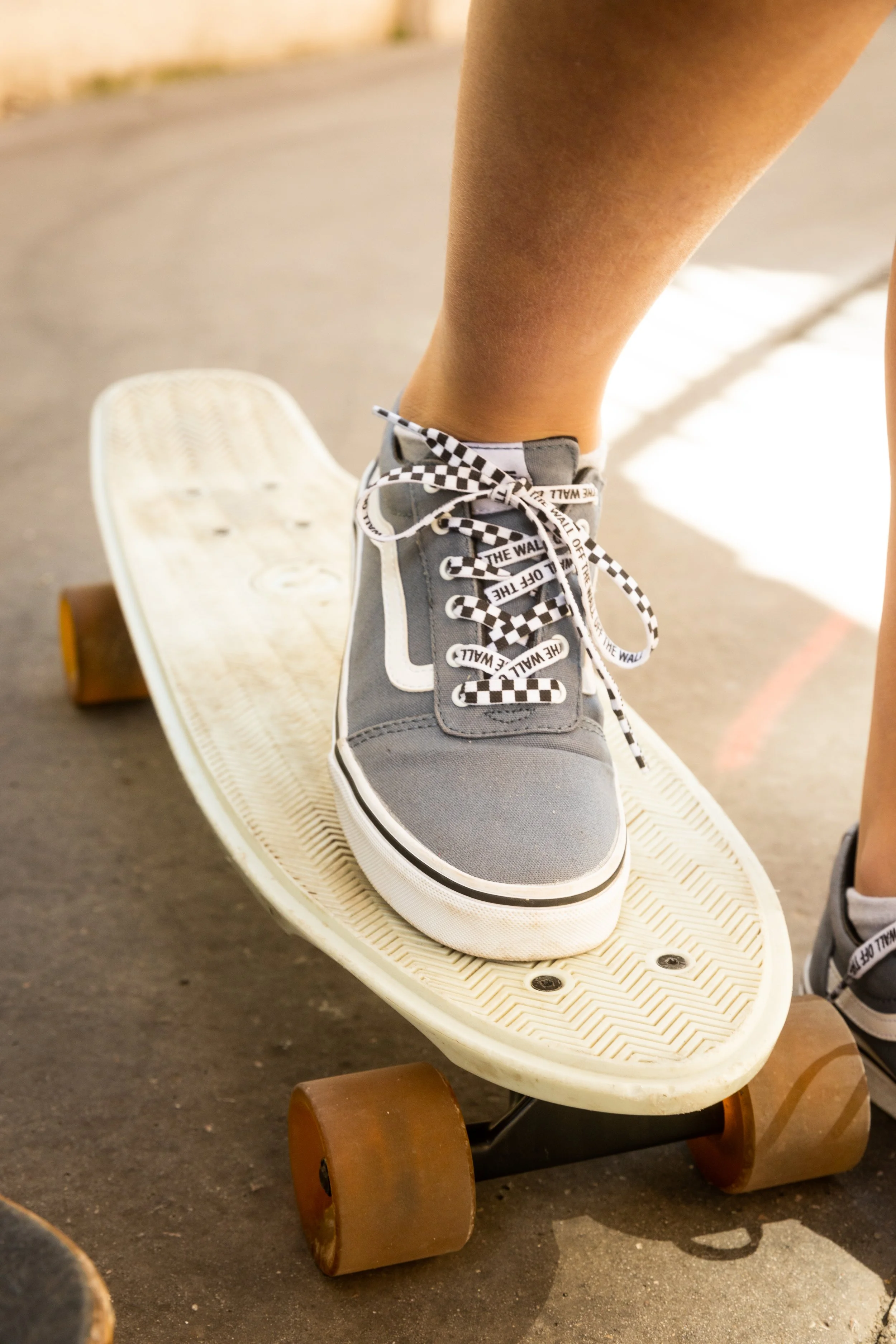 Un pied en skateboard portant une chaussure grise avec des lacets à motif de damier noir et blanc, sur un sol asphalté.