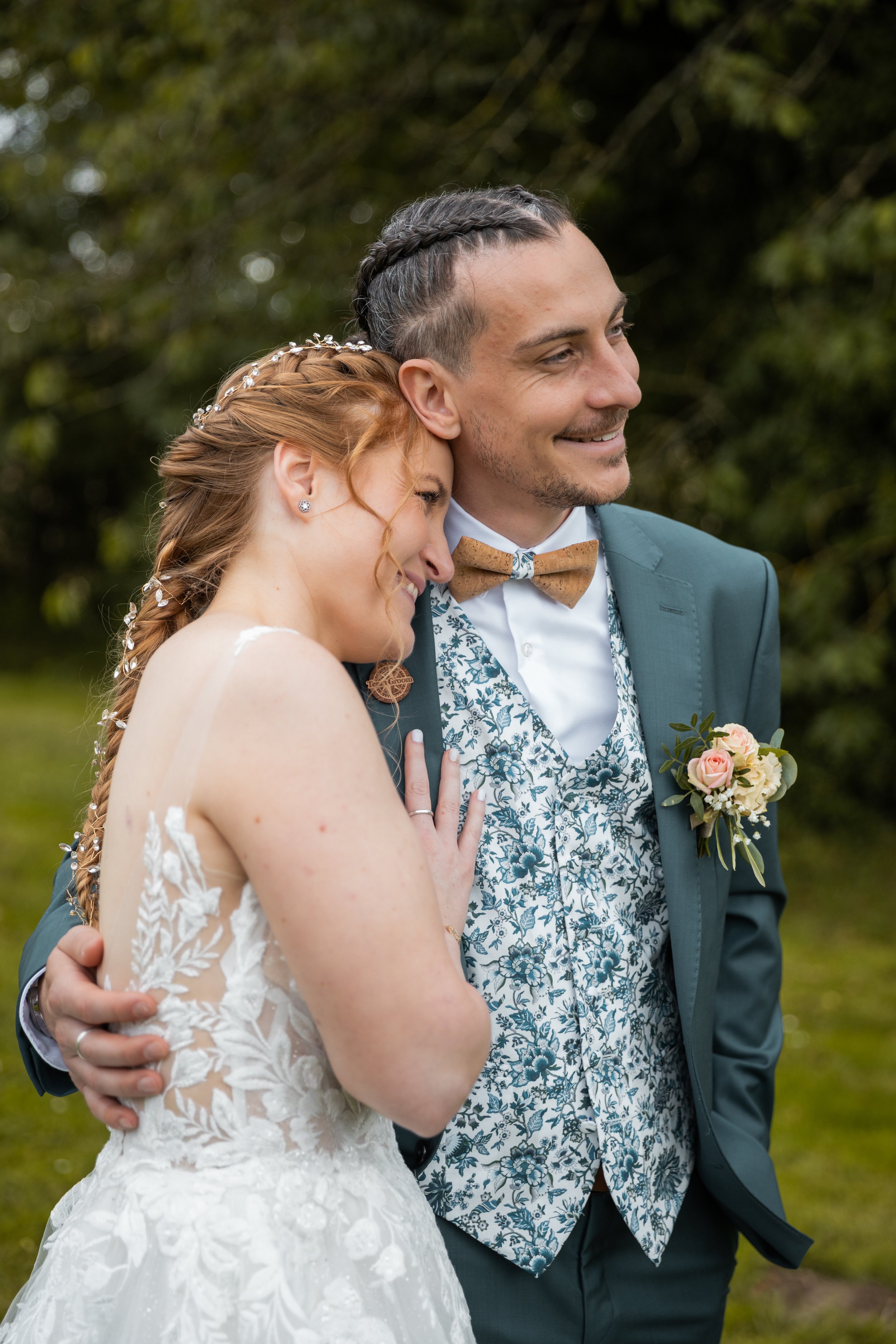 Un couple de mariés heureux partage un moment intime en plein air. La femme porte une robe de mariée blanche avec des détails floraux et une tresse ornée de petits fleurs. L'homme porte un costume bleu avec un gilet à motifs floraux, une chemise blan