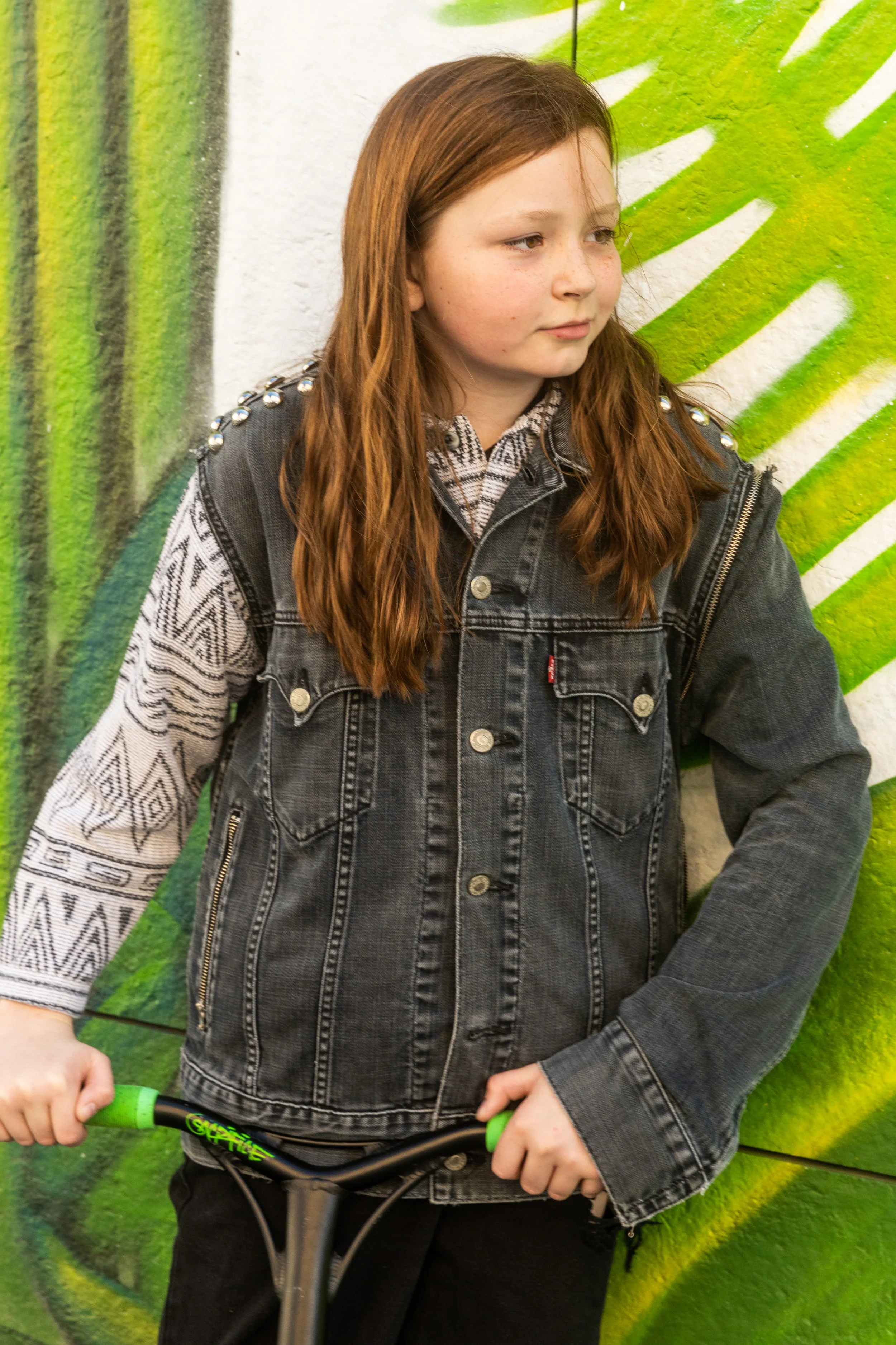 Jeune fille avec des cheveux roux, portant une veste en jean noir avec des épaulettes ornées de perles et une chemise à motifs, tenant un scooter, devant un mur peint avec un dessin vert et blanc.