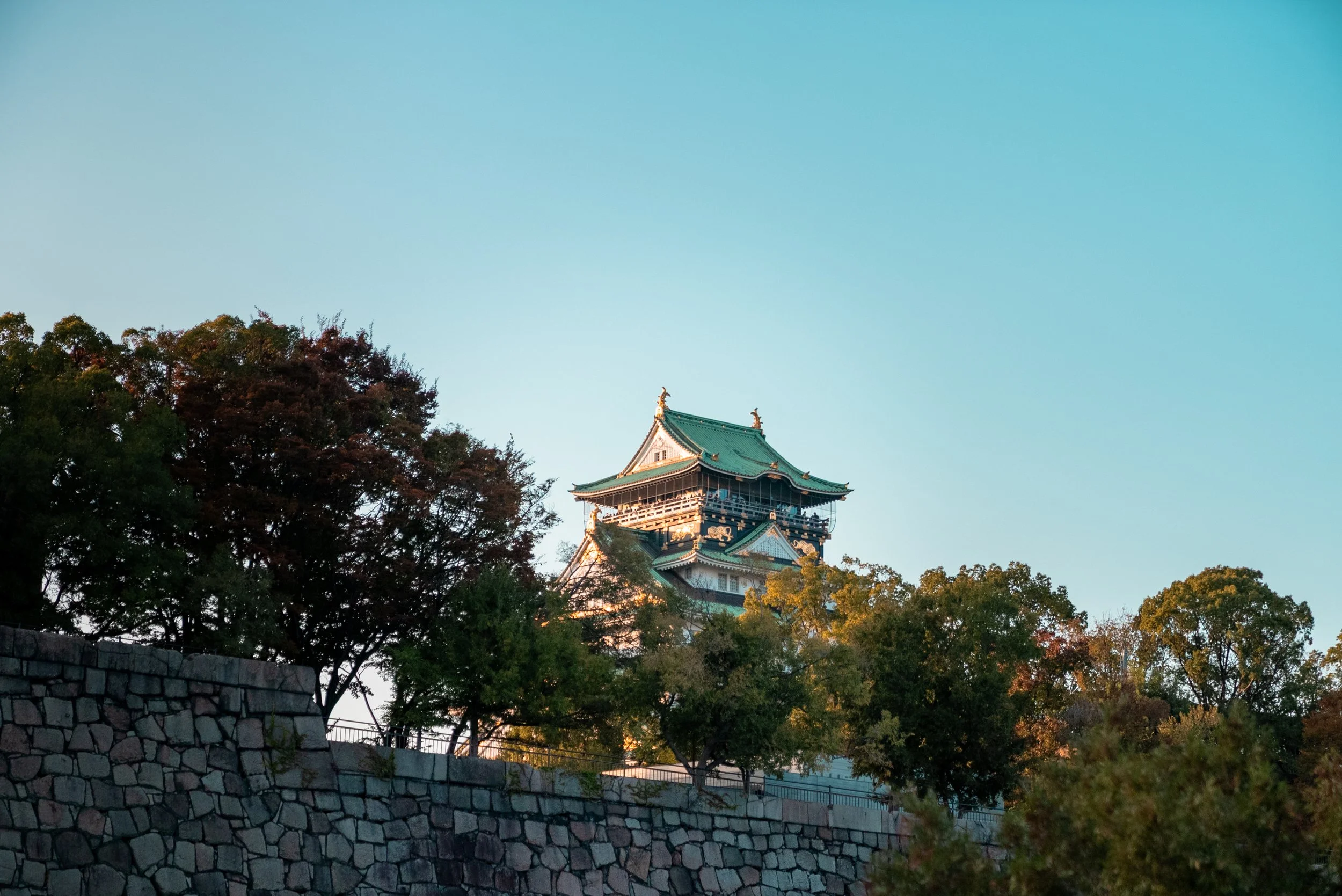Château japonais traditionnel surplombant une terrasse en pierre, entouré d'arbres en automne, ciel clair