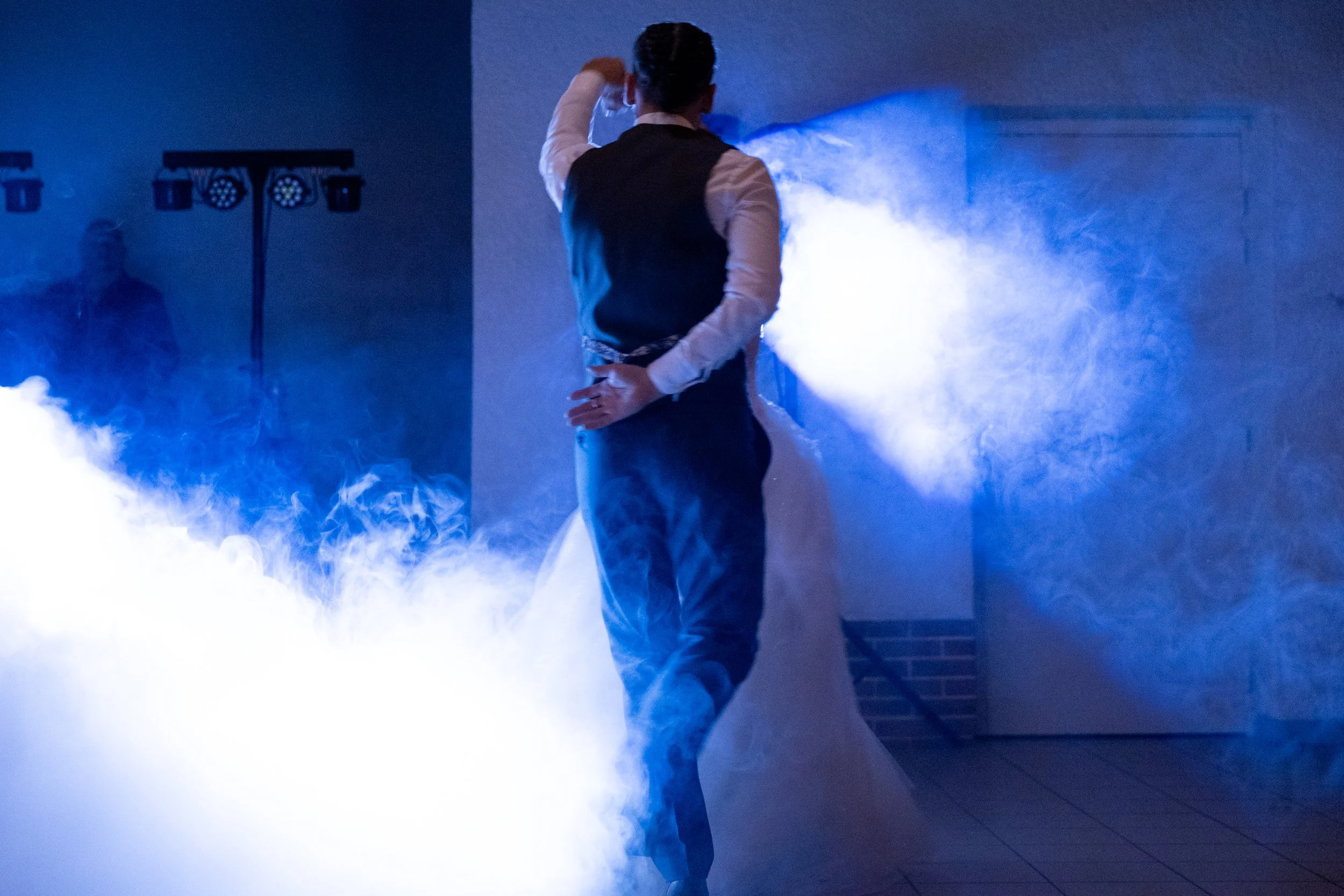 Un couple danse lors de leur mariage, entouré de fumée bleue, dans une pièce avec un mur blanc et un petit porte en bois.