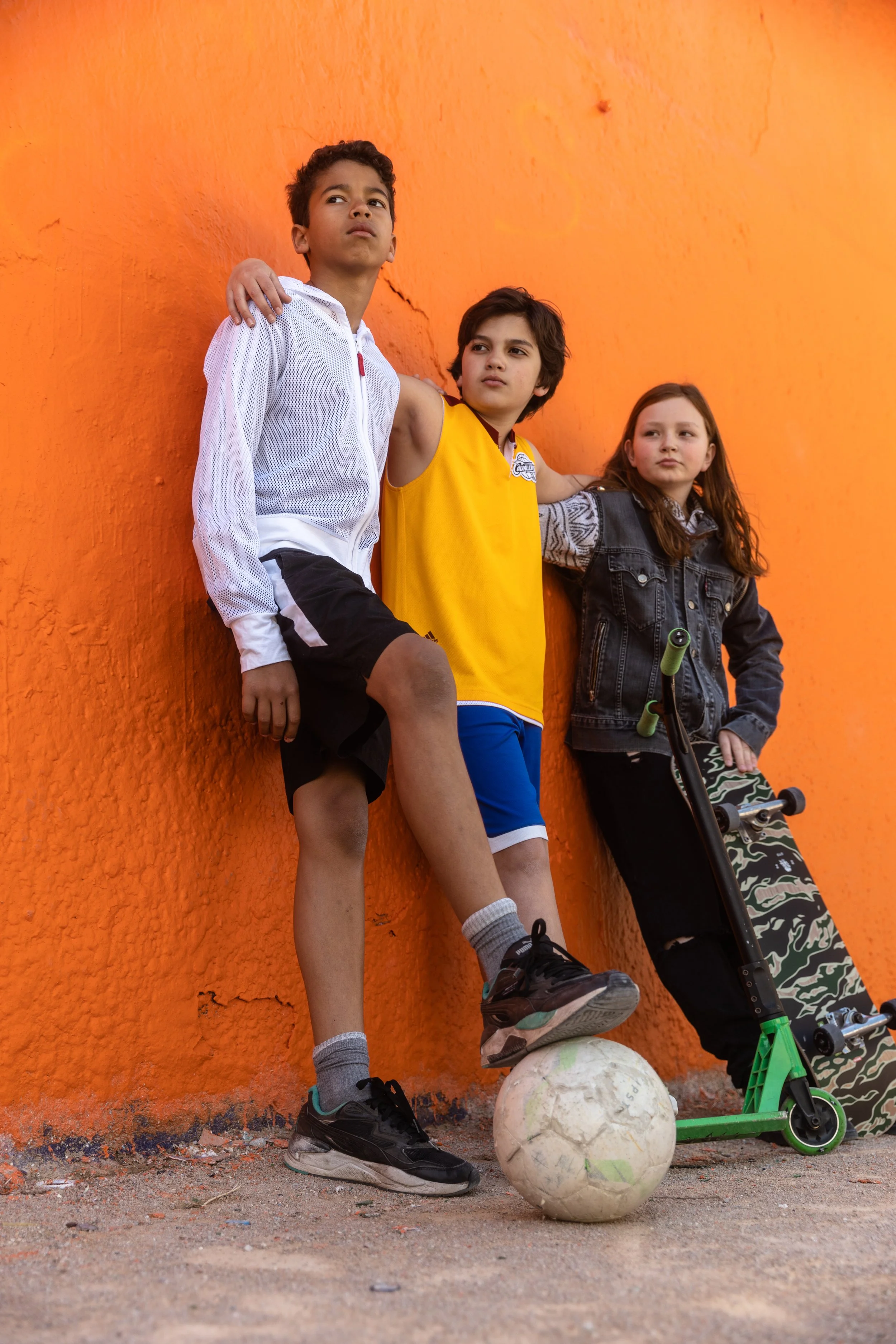Trois enfants posant contre un mur orange, un avec un ballon de football, un autre avec une planche à roulettes.