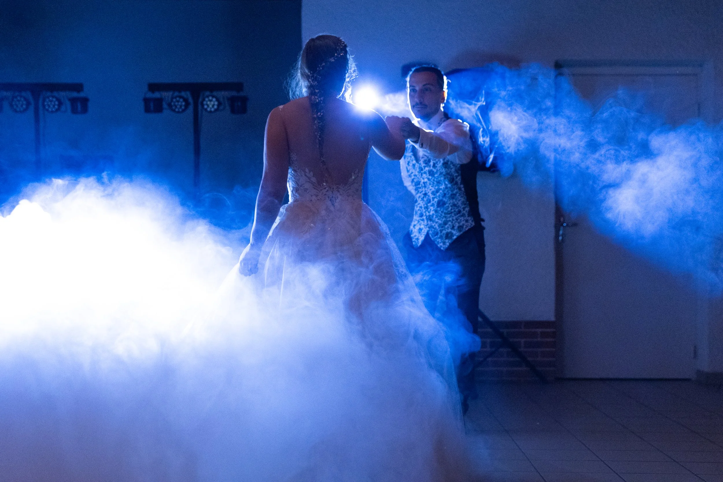 Un couple danse lors d'une cérémonie, entouré de fumée bleue, avec un éclairage lumineux derrière eux.