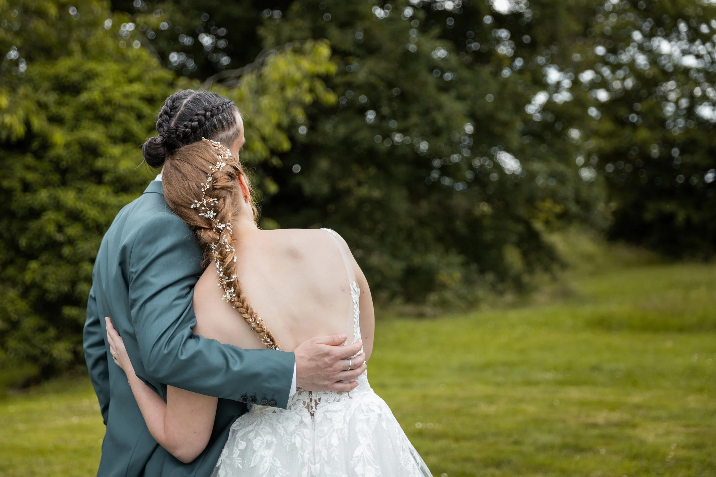 Un couple de mariés embrassant dans un parc vert, le marié porte un costume bleu vert, la mariée porte une robe de mariage blanche avec dos nu et une coiffure tressée ornée de bijoux floraux.