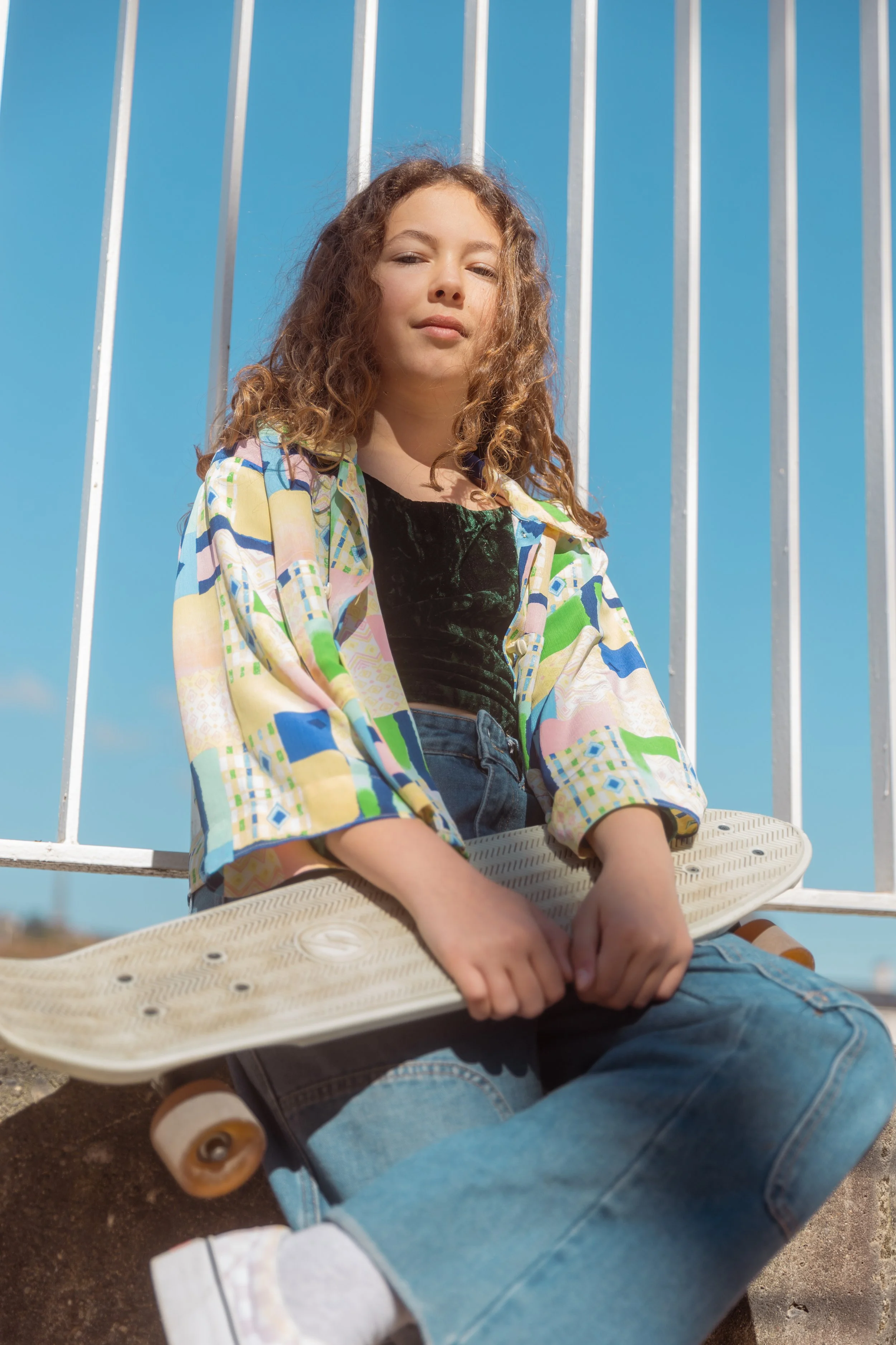 Jeune fille assise avec une planche à roulettes contre une barrière en métal, portant un chemisier coloré et un haut noir, sous un ciel bleu clair.