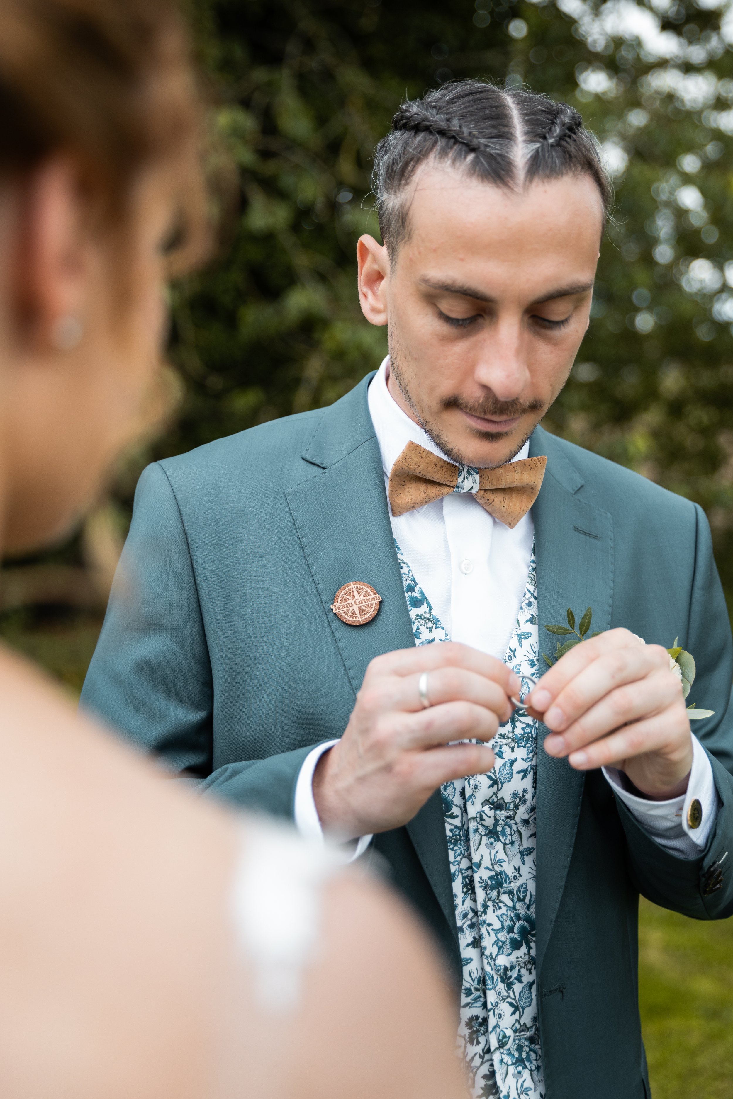 Un homme habillé en costume vert avec un nœud papillon en bois, attachant quelque chose lors d'un mariage ou d'une cérémonie en plein air.