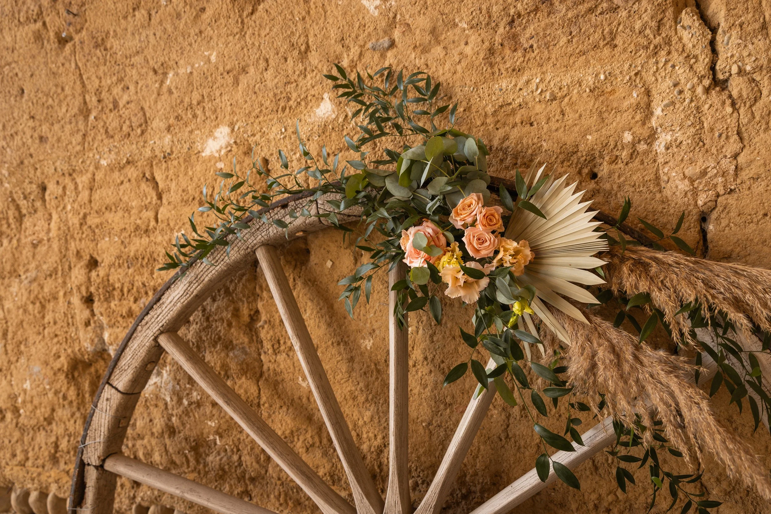 Une décoration florale suspendue sur une roue en bois contre un mur en terre cuite.
