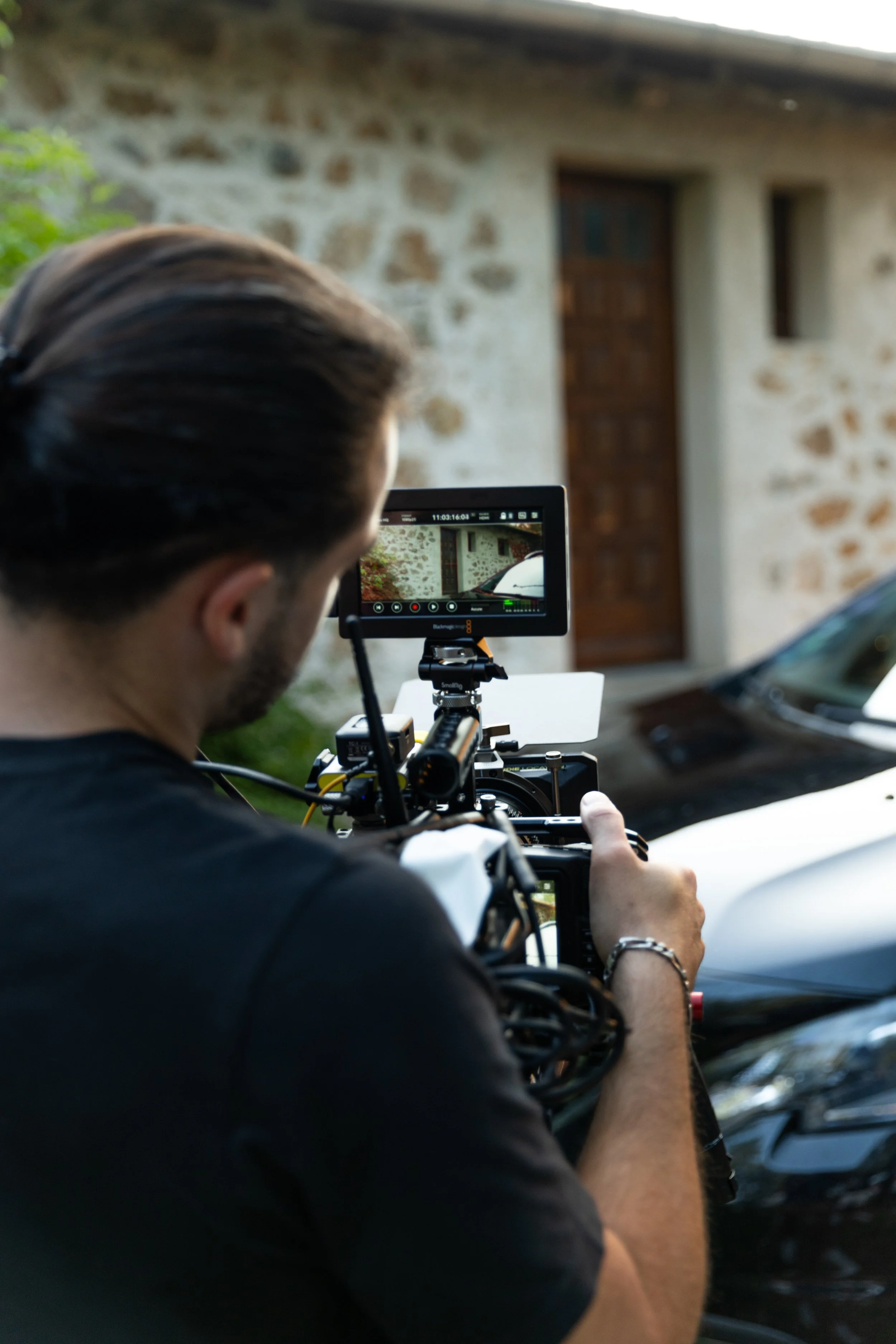Un réalisateur ou photographe en train de filmer ou prendre une photo d'une scène devant une maison en pierre, avec une voiture noire à côté.