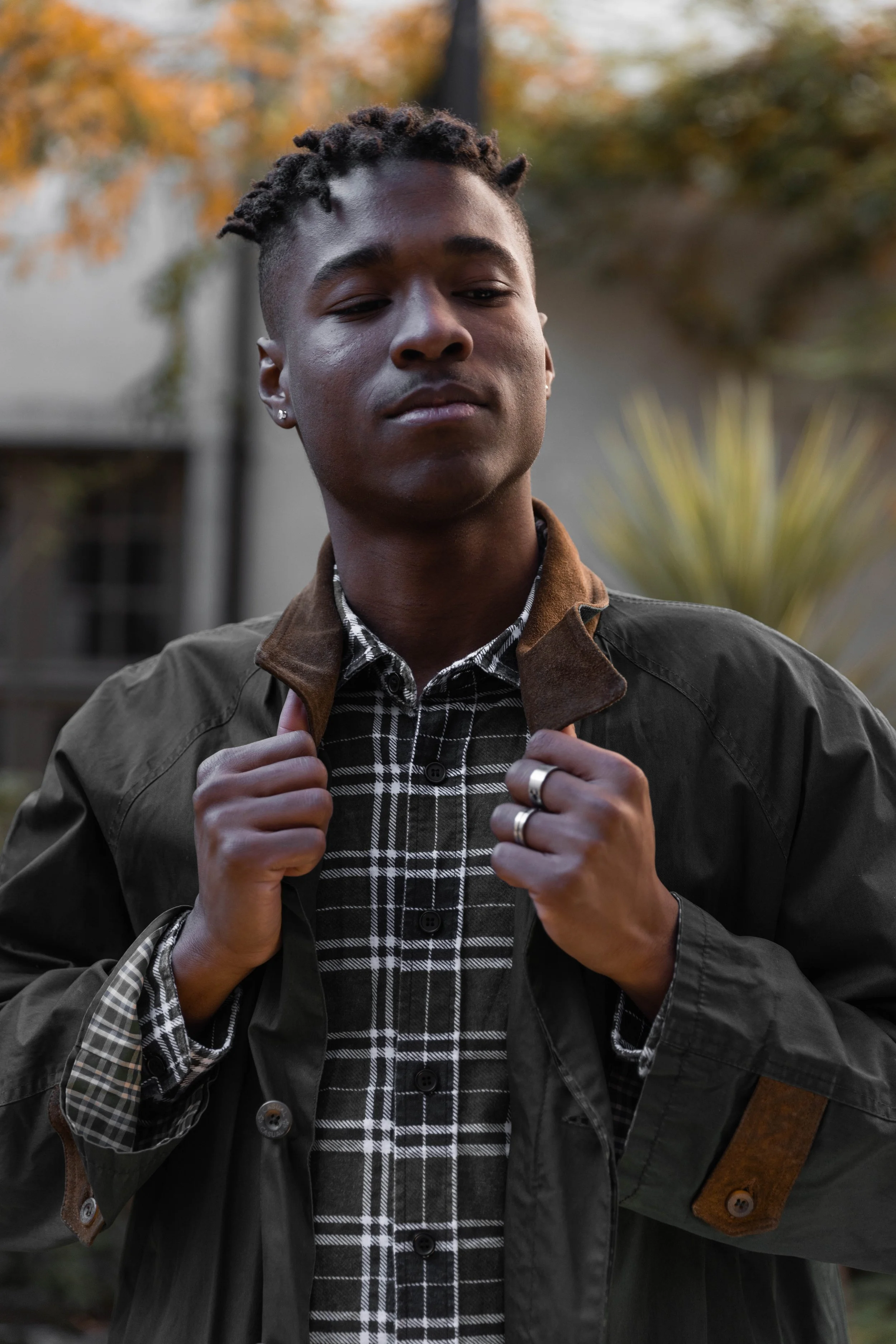 Jeune homme avec des dreadlocks, portant une veste et une chemise à carreaux, se tenant en extérieur avec un arrière-plan de feuillage automnal.