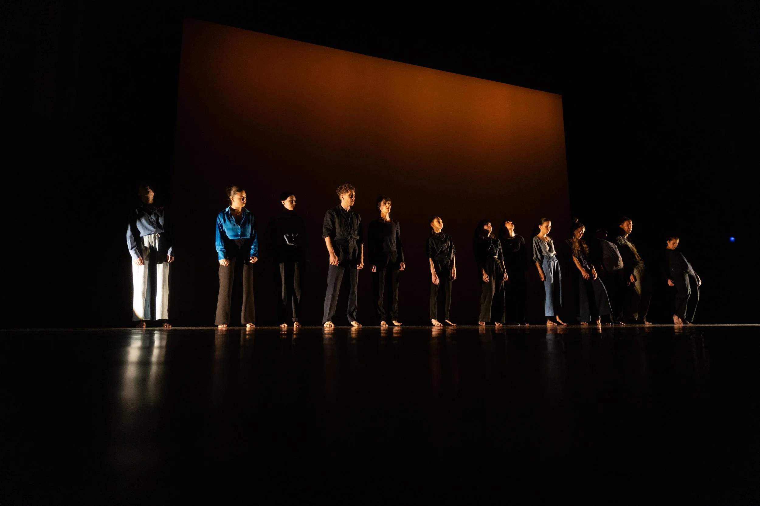 Groupe de personnes debout en ligne sur scène pendant une performance ou une répétition, fond sombre avec une lumière douce sur eux.