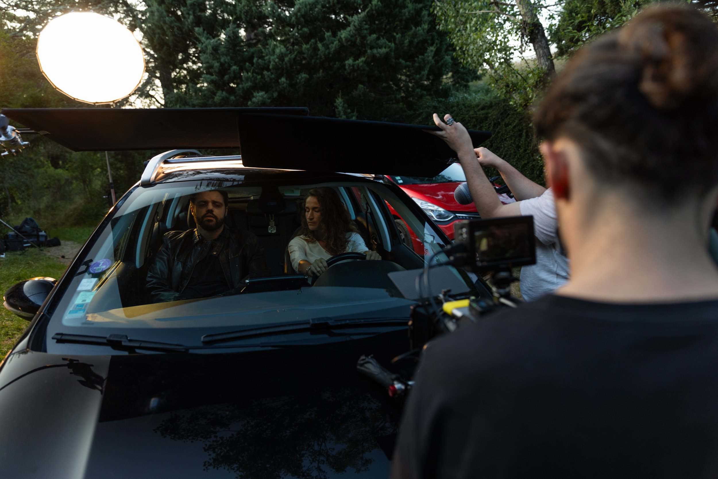 Tournage d'une scène de film ou de vidéo avec deux acteurs dans une voiture, plusieurs personnes en dehors préparant l'éclairage et le matériel de tournage, dans un environnement extérieur avec des arbres.