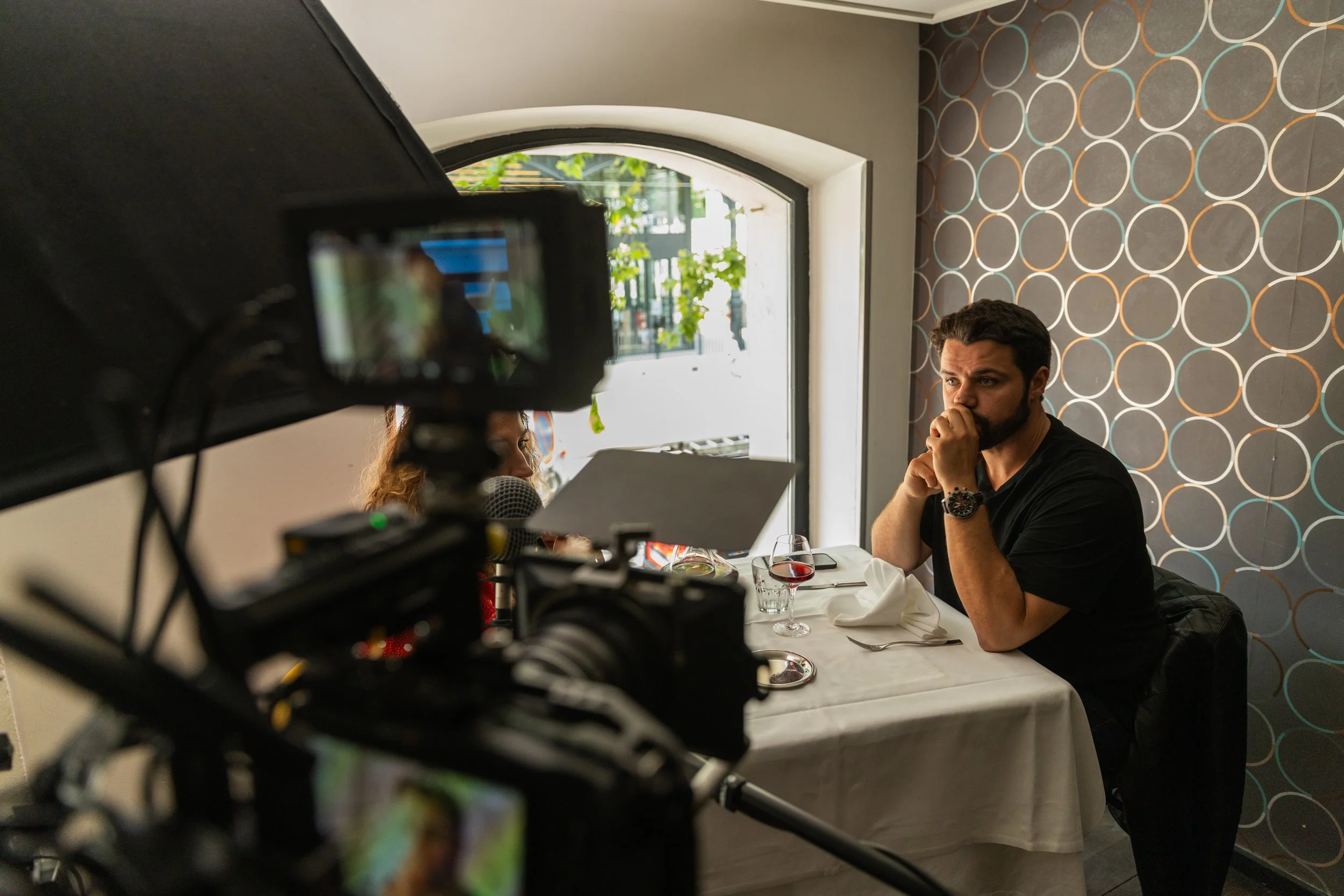 Une personne à une table de restaurant regarde fixement un caméscope, tandis qu'une autre personne parle dans un microphone, lors du tournage d'une scène, avec un mur à motifs circulaires en arrière-plan.