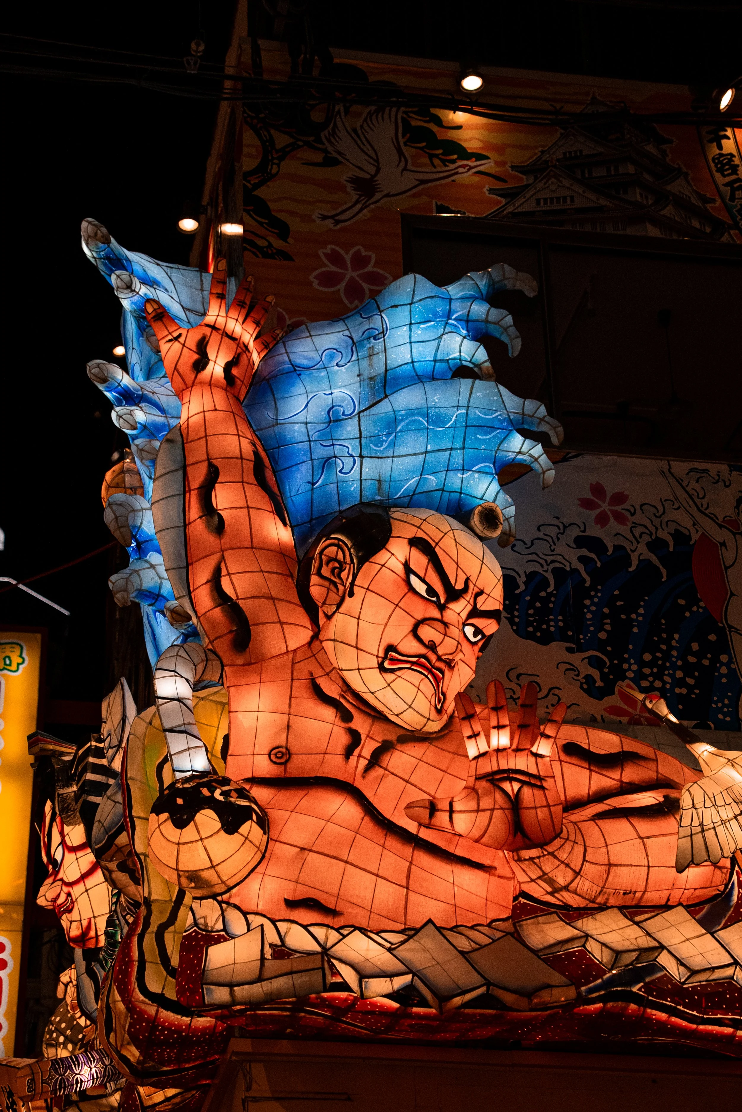 Une grande lanterne en forme de samouraï avec une expression féroce et un kimono rouge, tenant une arme, dans une parade de lanternes japonaises nocturnes.