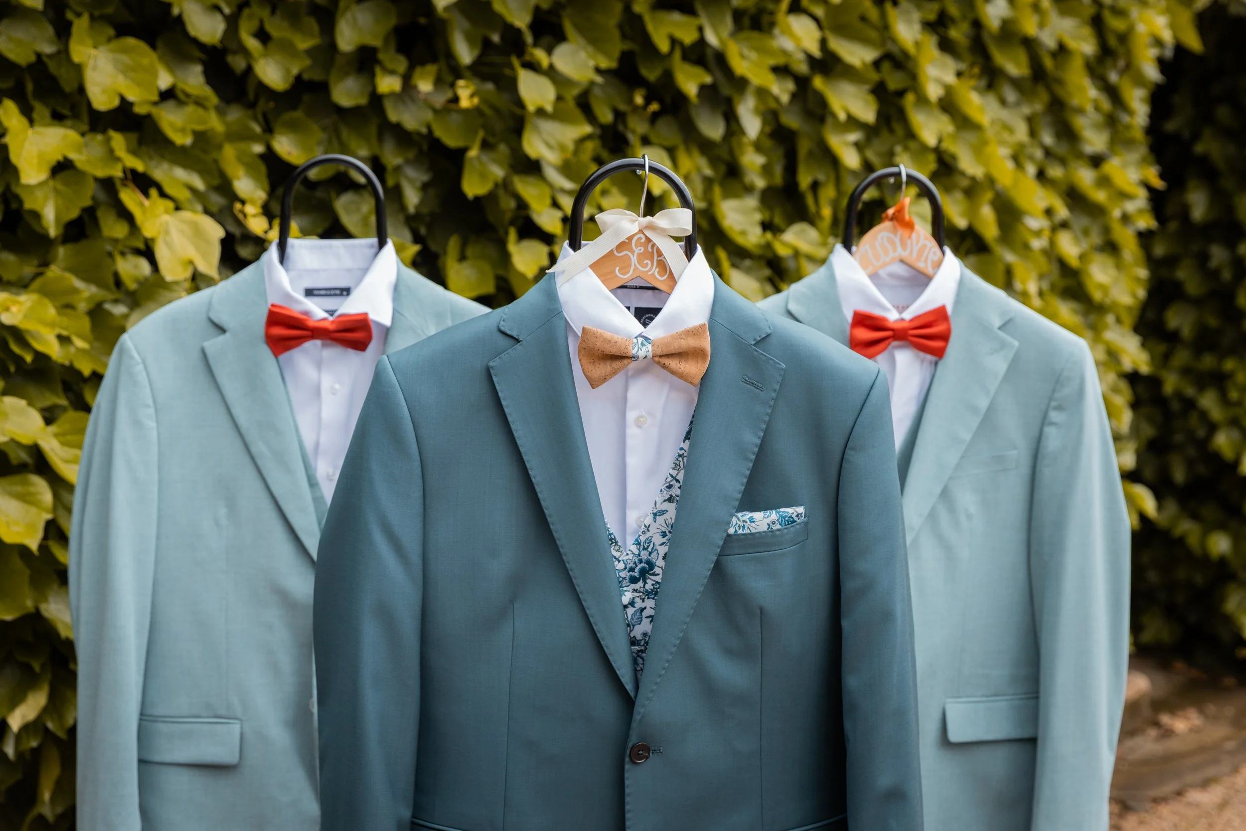 Costumes de mariage avec costumes bleus et nœuds papillons rouges ou en bois, suspendus contre un mur de feuillage vert.