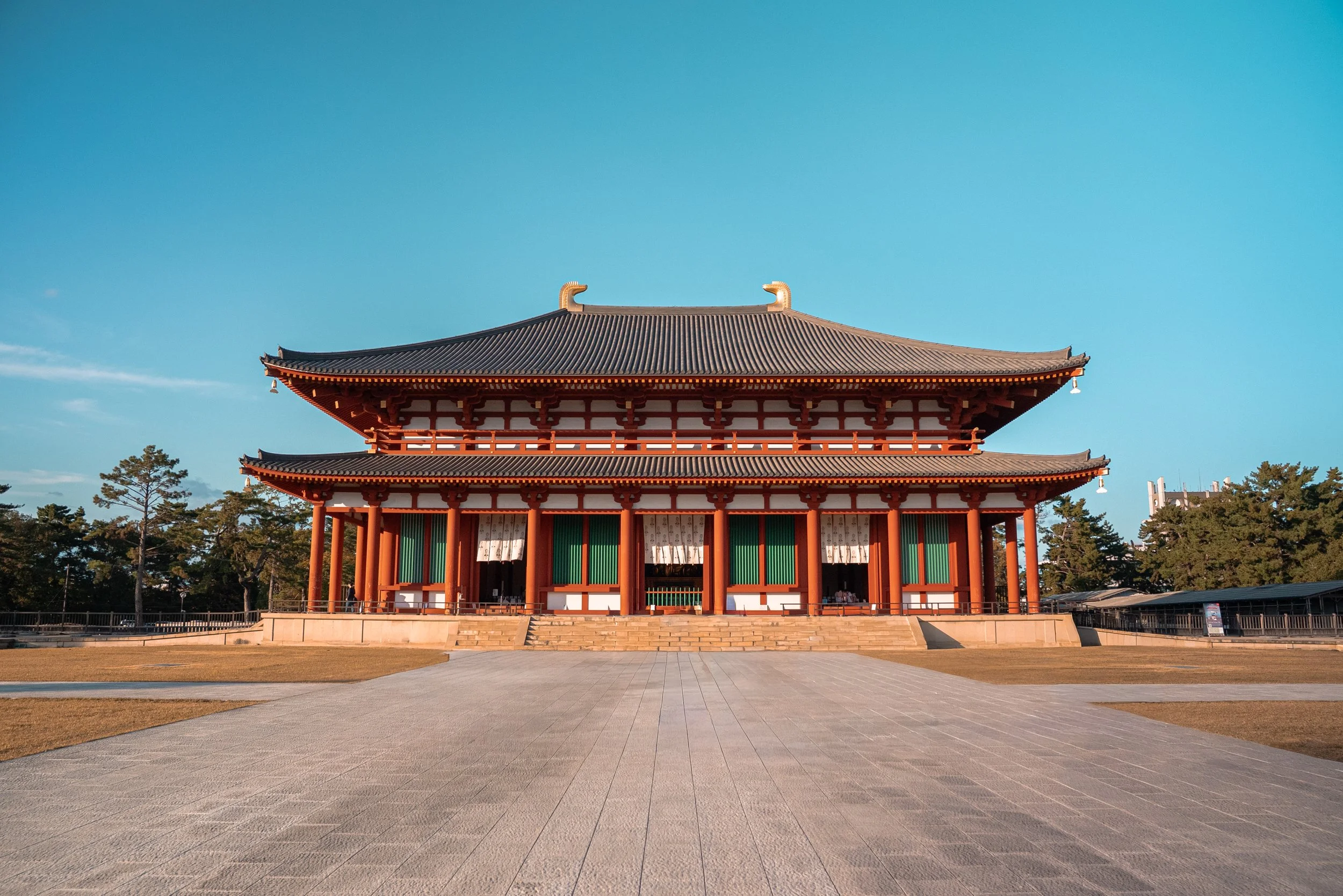 Un bâtiment traditionnel japonais avec un toit à deux niveaux, échafaudages en bois rouges et entouré d'arbres, sous un ciel clair.