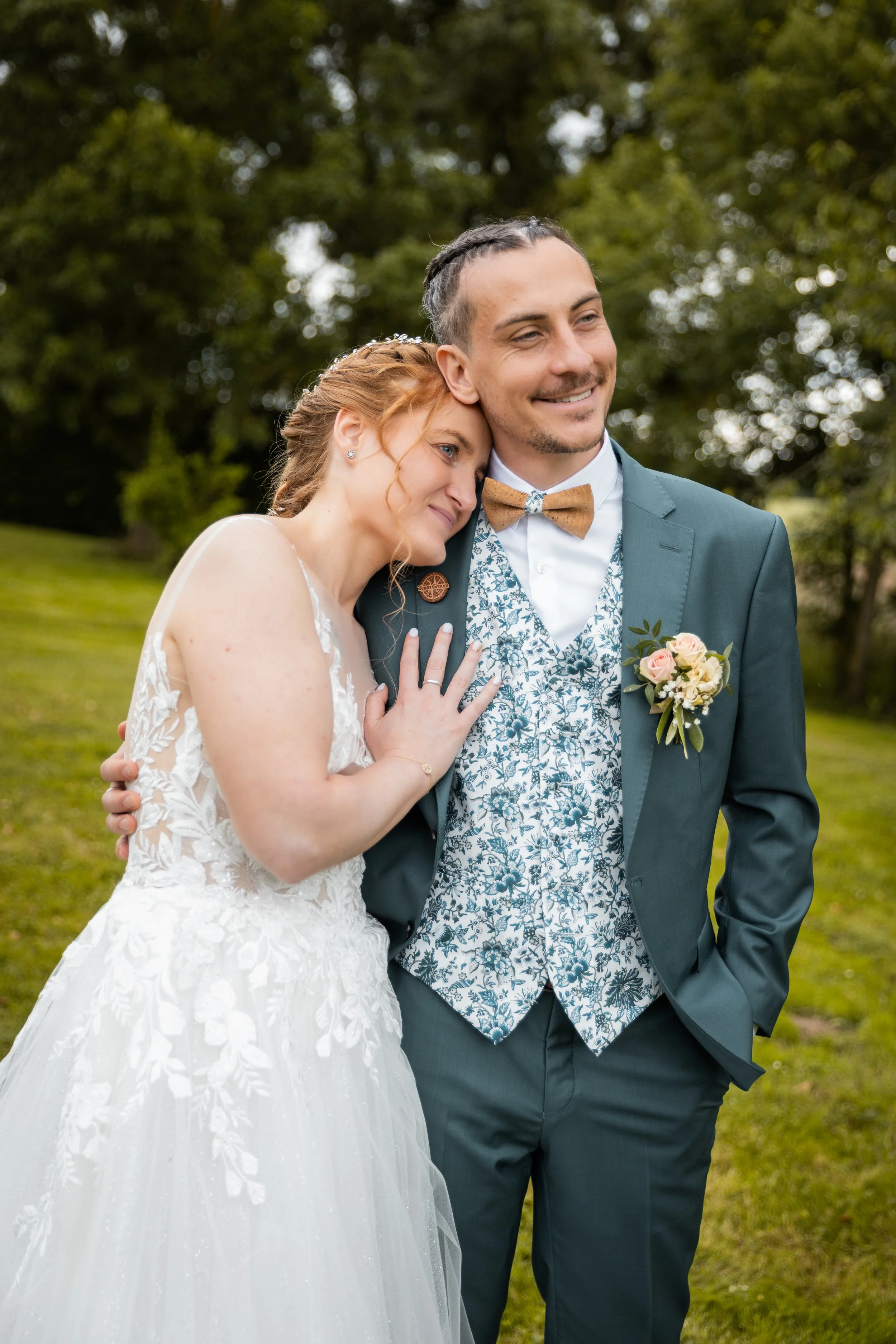 Un couple marié souriant, la femme en robe de mariée blanche et le homme en costume avec une veste à motif floral, dans un parc verdoyant.