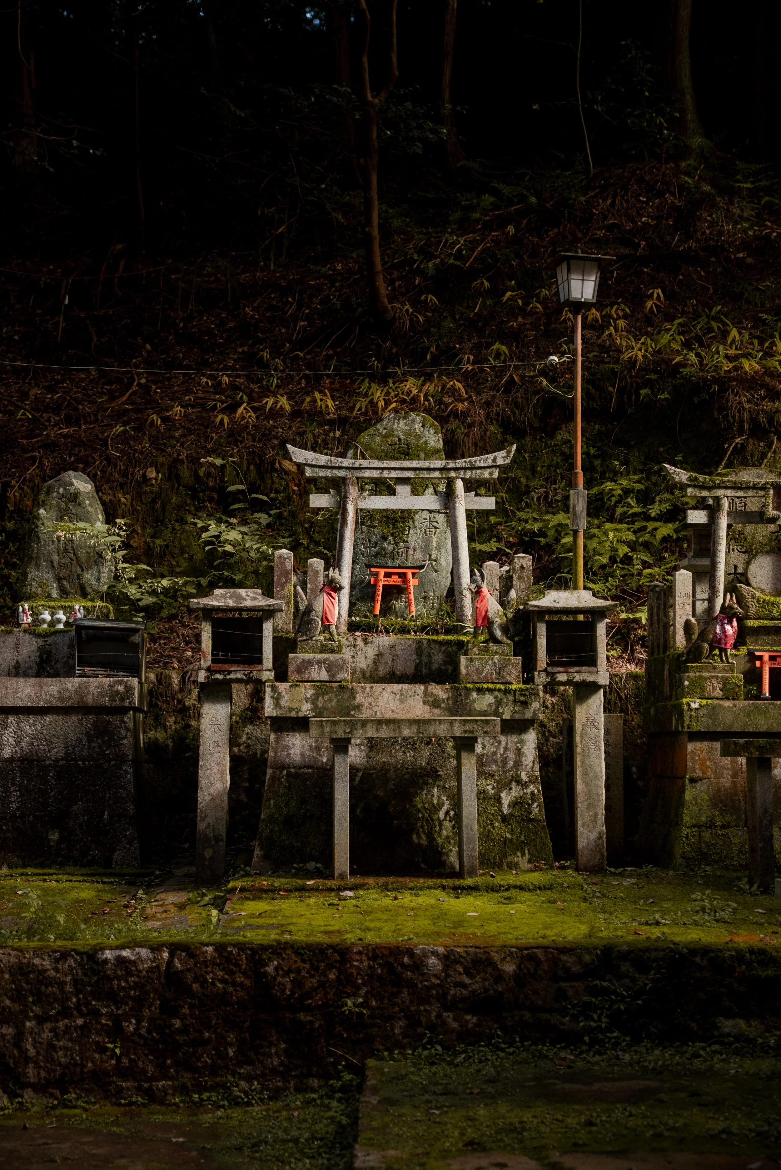 Des petits sanctuaires shintoïstes en pierre, entourés de lanternes, dans un environnement forestier, avec des mousses vertes sur le sol et des statues de renards porteurs de vêtements rouges.