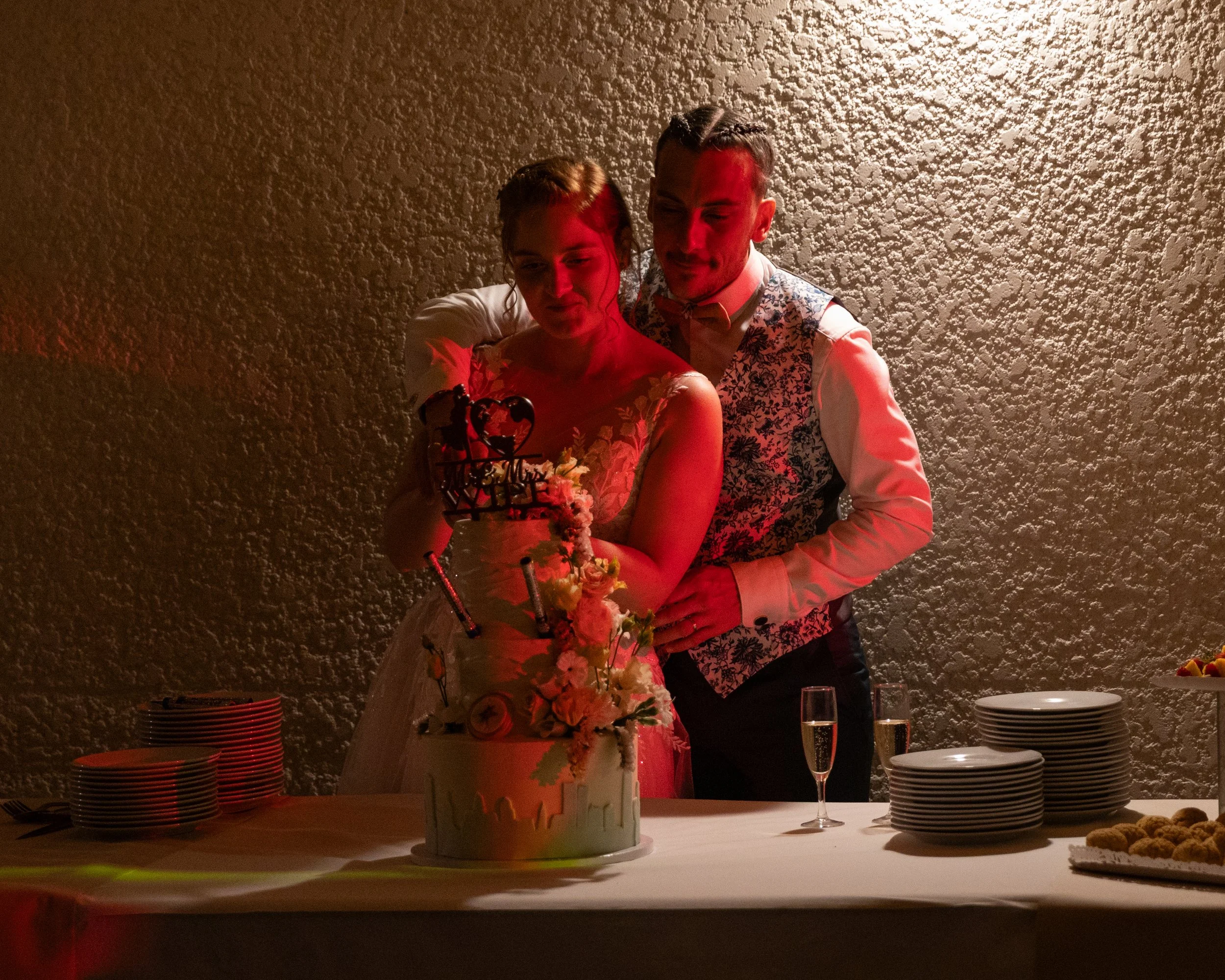 Un couple célèbre leur mariage avec un gâteau au centre d'une table. La mariée porte une robe blanche et le marié un veston à motifs. Il y a des assiettes, des couverts, des verres de champagne et des plats de nourriture sur la table. La décoration e
