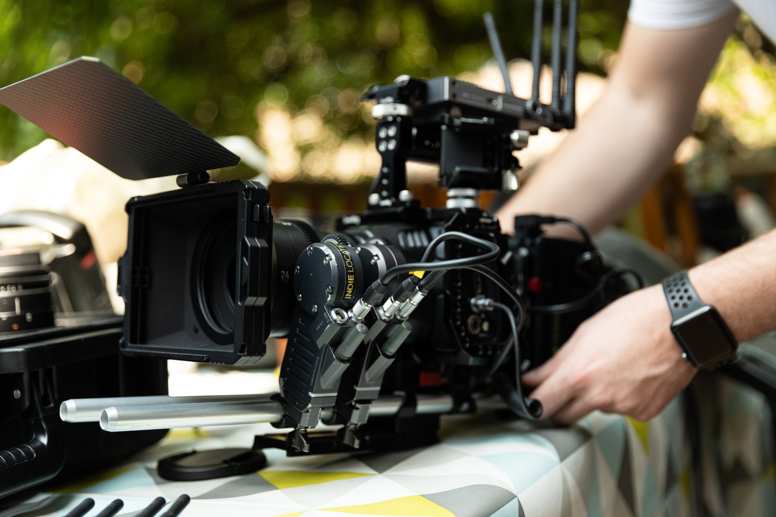 Un opérateur manipulant une caméra professionnelle équipée d'accessoires et de câbles, posé sur une table extérieure en journée.