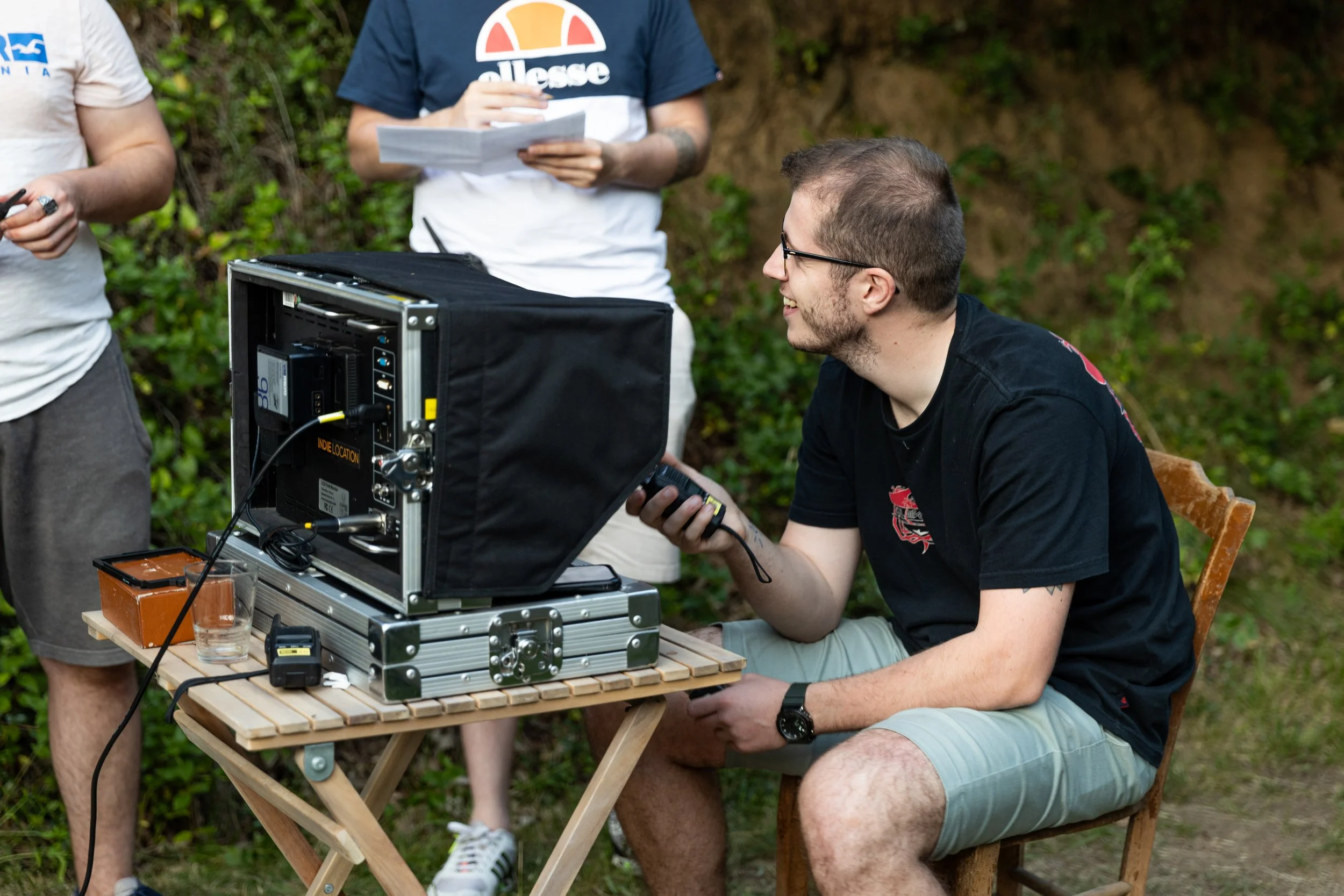 Un homme souriant assis à une table en extérieur, utilisant une télécommande, avec un ordinateur portable ou un équipement technique placé devant lui. Deux autres personnes se tiennent à côté, une tenant une feuille, tous dans un environnement nature