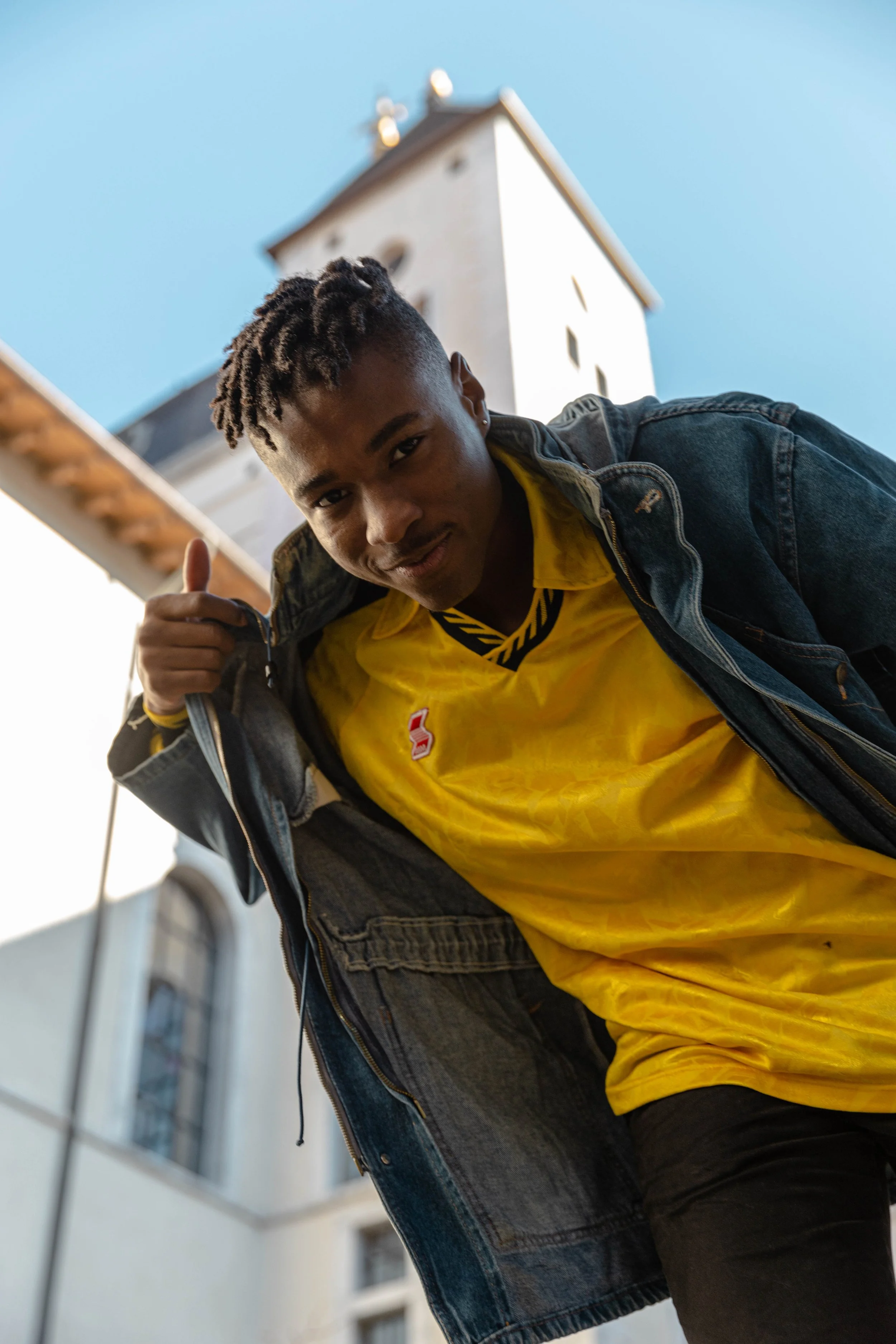 Jeune homme souriant, portant une veste en jean et un t-shirt jaune, faisant un geste de thumbs-up, avec une église en arrière-plan, ciel clair.